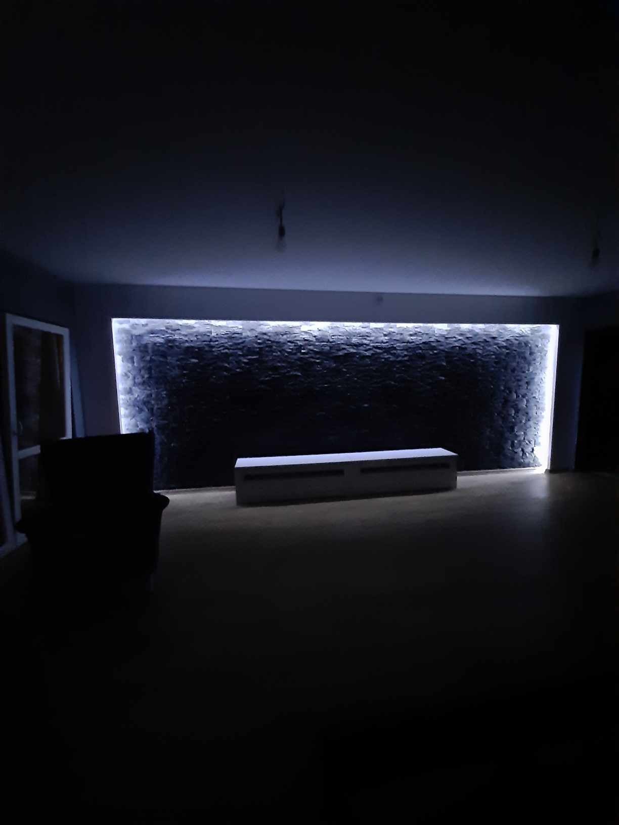 Ściana z dekoracyjnego kamienia podświetlona ledami w ciemnym pomieszczeniu, widoczna szafka RTV i fragment okna po lewej.