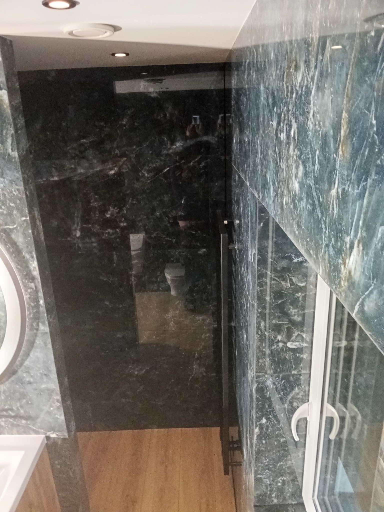 Luksusowa łazienka z marmurowymi ścianami w odcieniach szarości i zieleni, kabina prysznicowa ze szklanymi drzwiami, drewniana podłoga i okrągłe lustro.