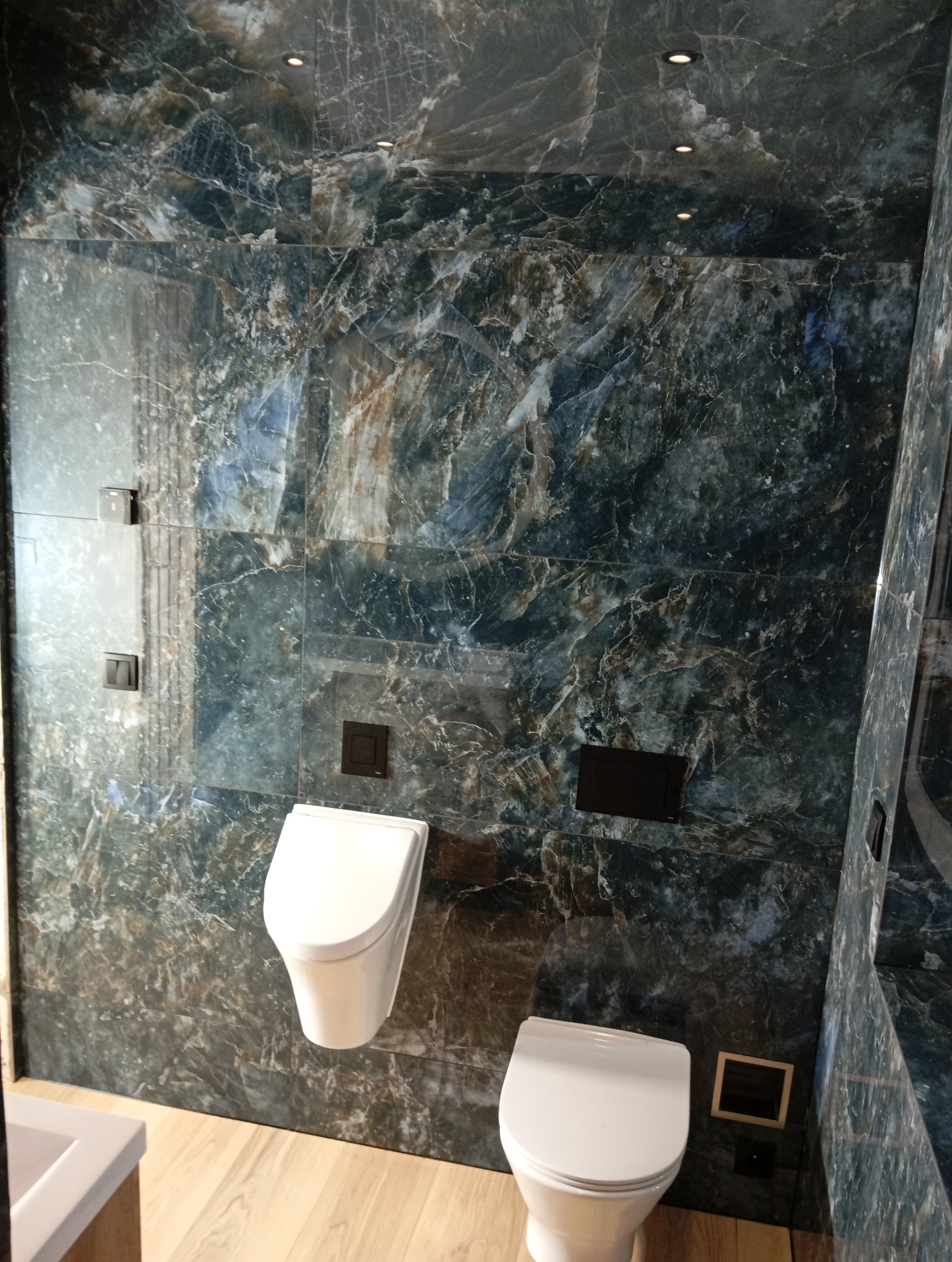 Elegancka łazienka z marmurowymi płytkami w odcieniach granatu i brązu. Wiszący bidet i toaleta, drewniana podłoga, minimalistyczne oświetlenie sufitowe.
