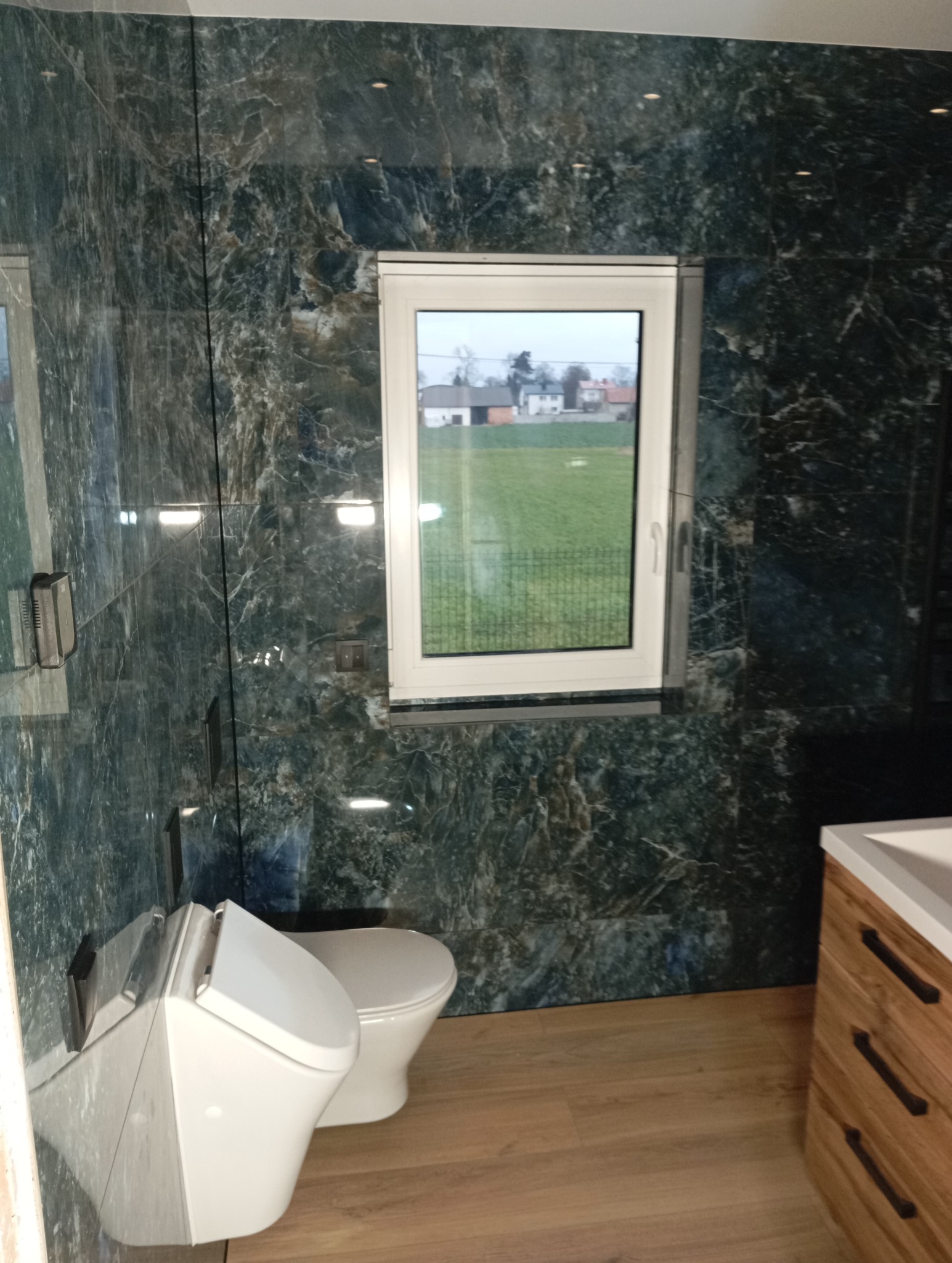 Elegancka łazienka z marmurowymi ścianami, jasną podłogą i oknem z widokiem na zieleń. Nowoczesna toaleta podwieszana i drewniana szafka z umywalką.