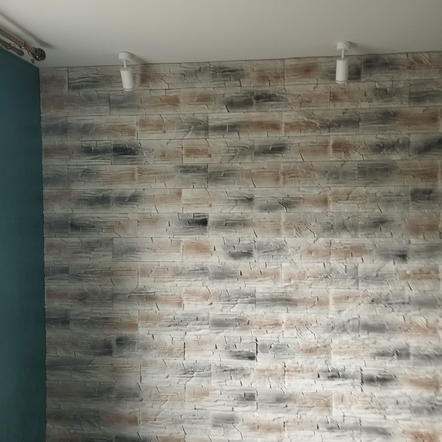 Ściana z dekoracyjnej okładziny imitującej kamień w odcieniach szarości i beżu, oświetlona dwoma reflektorami. Fragment turkusowej ściany po lewej.