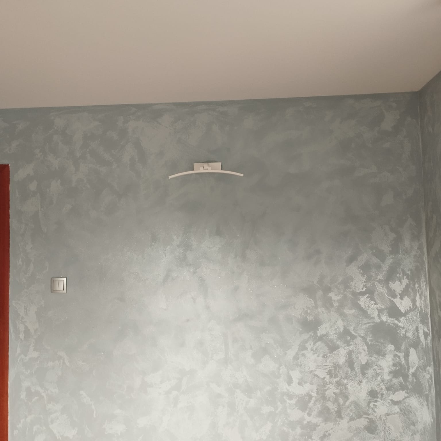 Ściana z efektem dekoracyjnym w odcieniach szarości, oświetlona lampą ścienną. Widoczny włącznik światła i fragment futryny drzwi. Subtelna tekstura na gładkiej powierzchni.