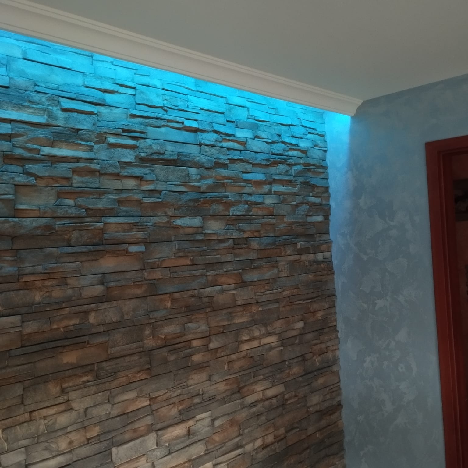 Ściana z dekoracyjnej cegły podświetlona niebieskim światłem LED, sąsiadująca z ścianą o fakturze w odcieniach błękitu i fragmentem futryny drzwi.
