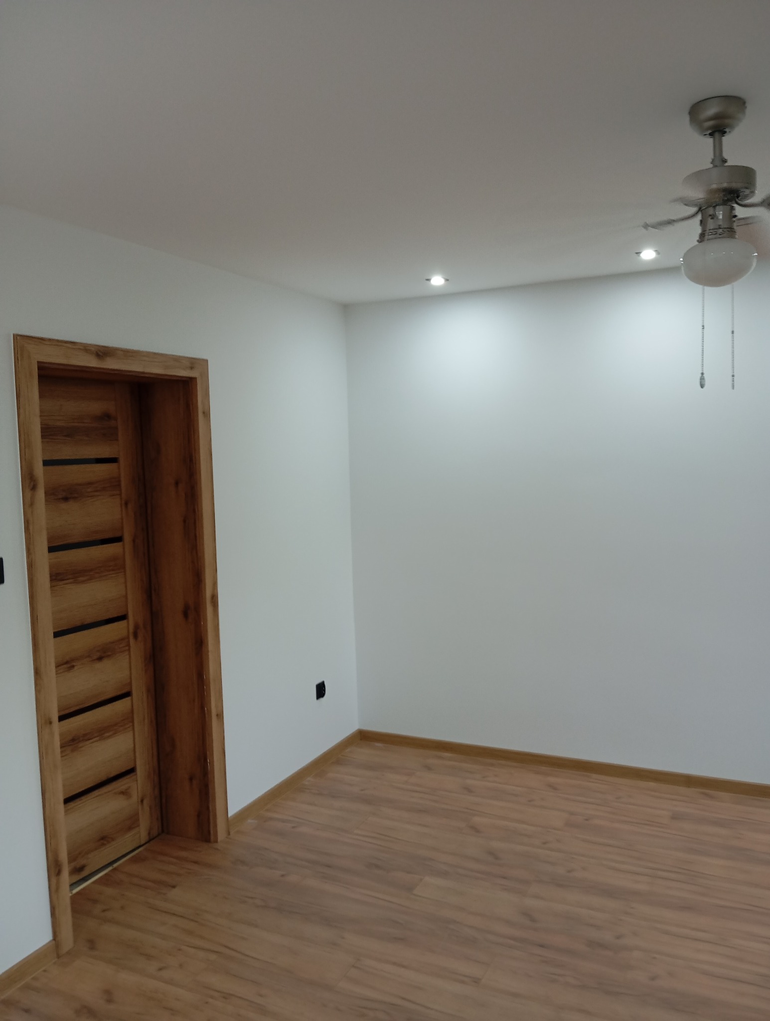 Wykończone wnętrze pokoju z drewnianą podłogą, białe ściany i drzwi z drewnianą ramą. Widoczny sufit z oświetleniem i wentylator.