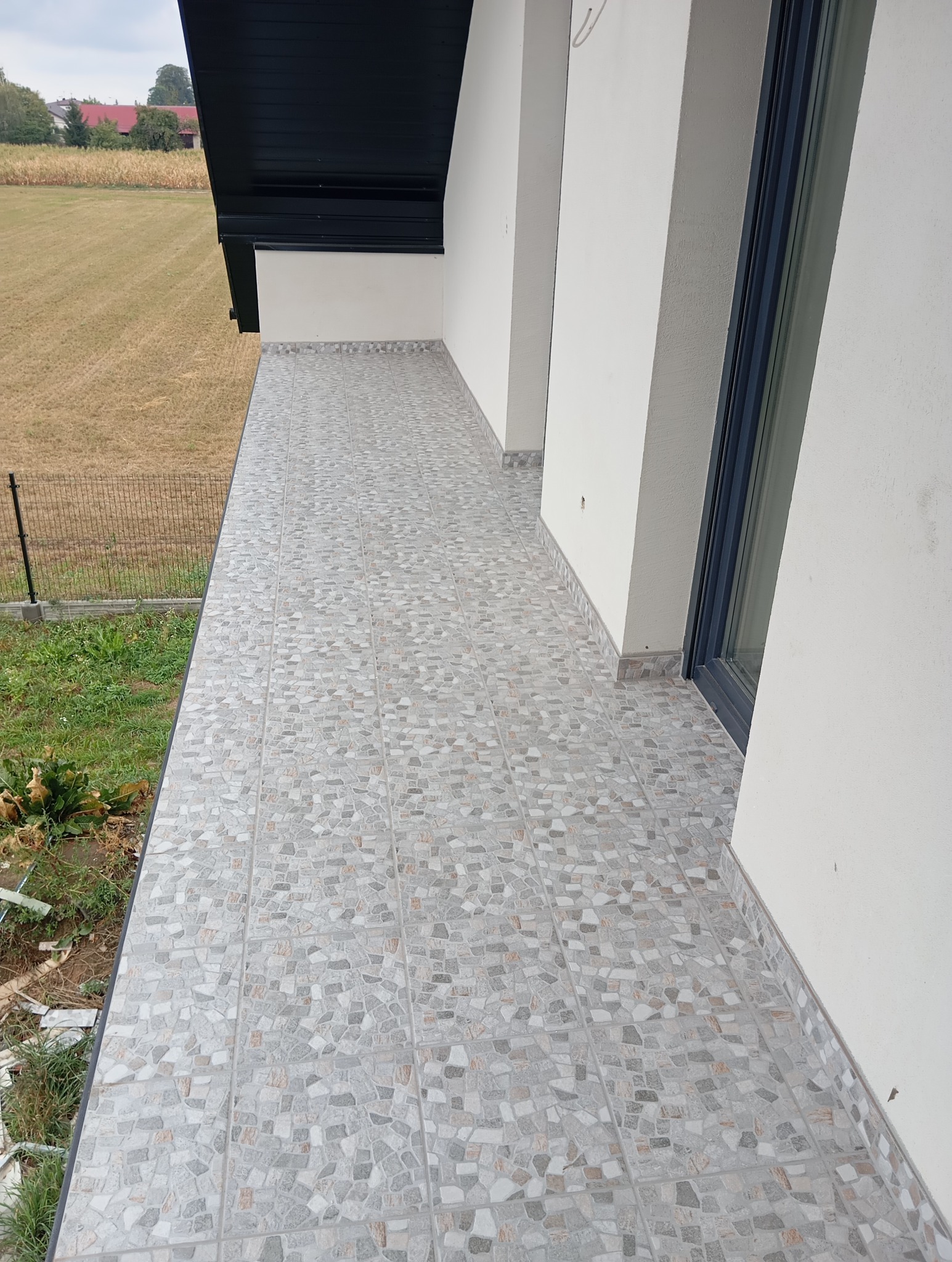 Wykończony balkon z szarą, mozaikową terakotą. Widok na pole przez czarną, metalową barierkę. Ściany białe, okno z ciemną ramą. Ukończona praca.