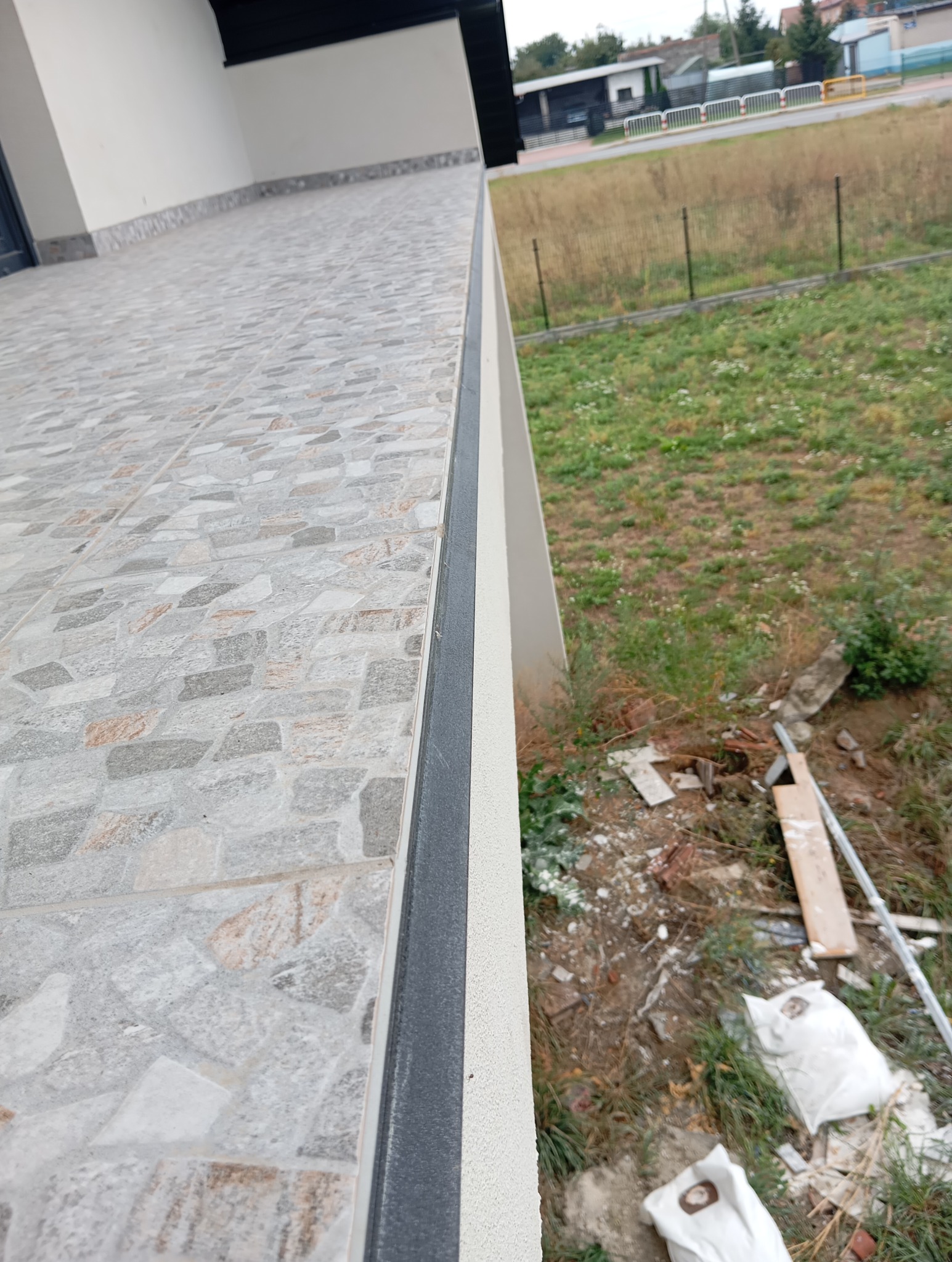 Wykończony balkon z szarymi płytkami o nieregularnym wzorze i czarną listwą wykończeniową. Widok z góry na otoczenie budynku.