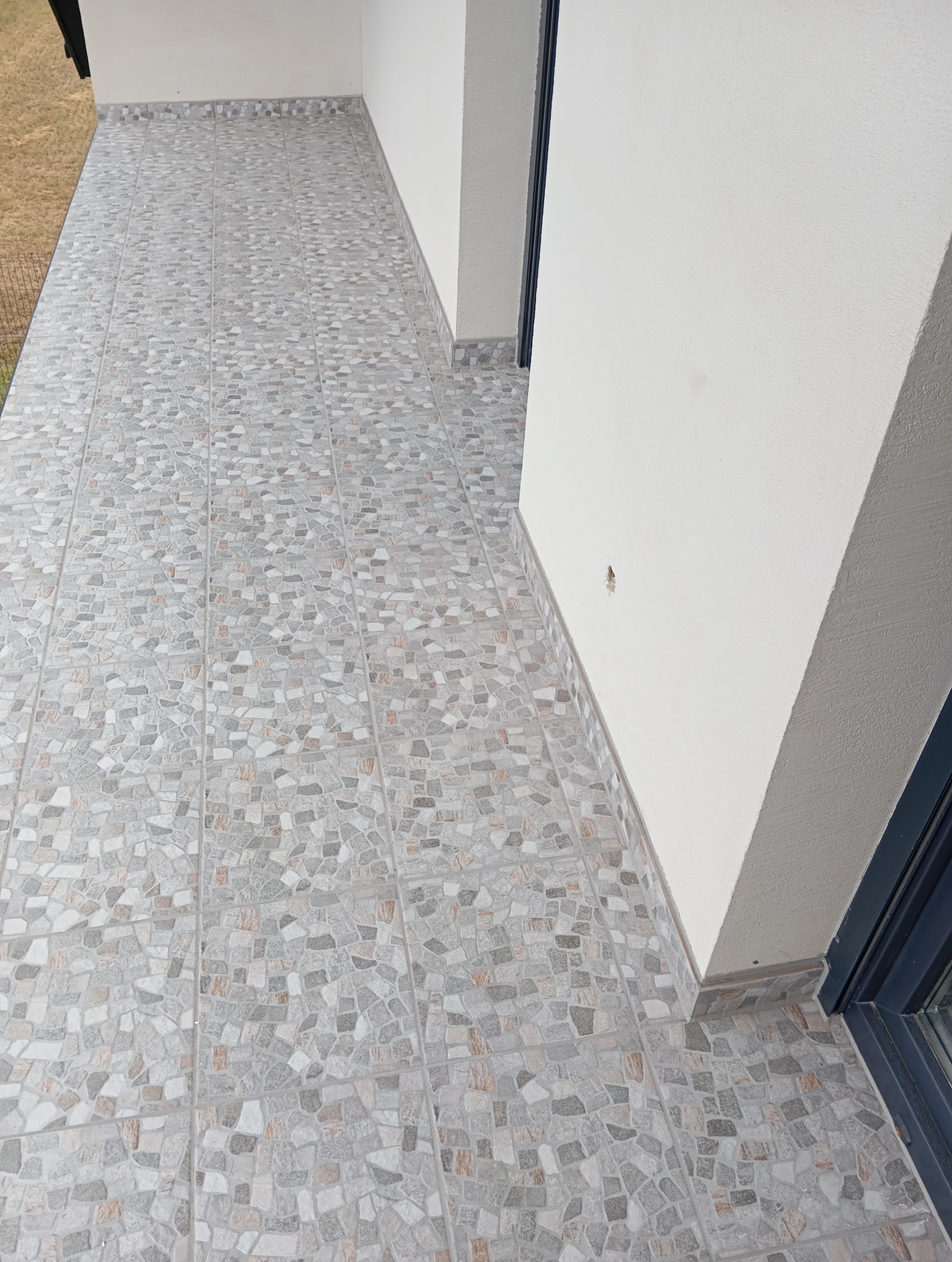 Wykończony taras z szarymi płytkami imitującymi kamień, z widoczną listwą przypodłogową i fragmentem ściany w tle. Płytki ułożone równo, z zachowaniem spójności wzoru.