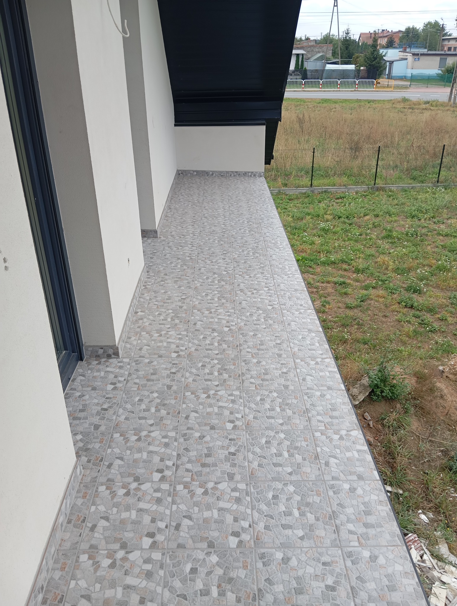 Wykończony balkon z szarymi płytkami imitującymi mozaikę. Widok na zewnątrz, z boku fragment ściany budynku z oknem. Ujęcie z góry, widać detale wykończenia.