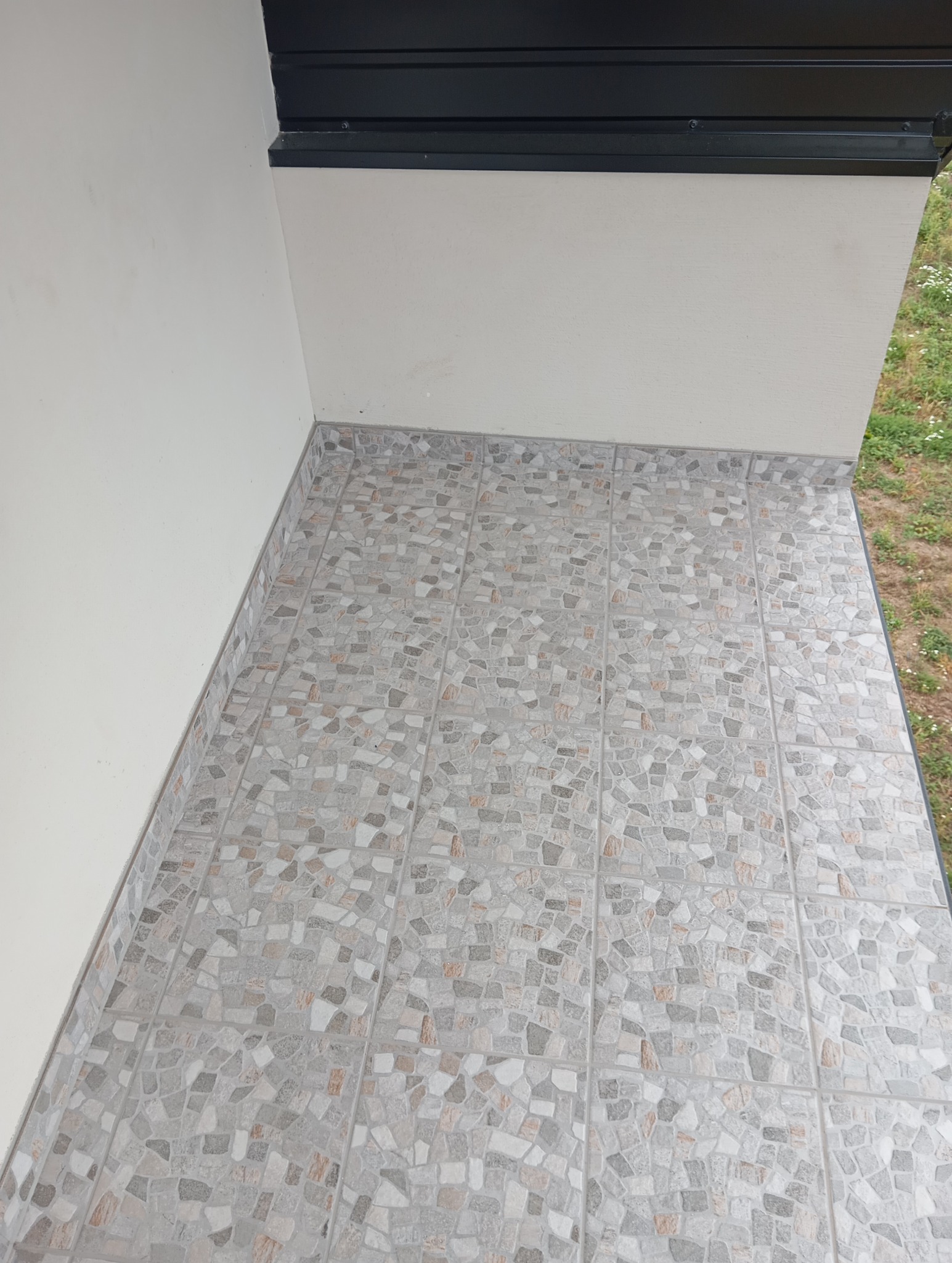 Wykończony balkon z mozaikową terakotą w odcieniach szarości i beżu, widok z góry, ściana w jasnym odcieniu, czarne wykończenie sufitu.