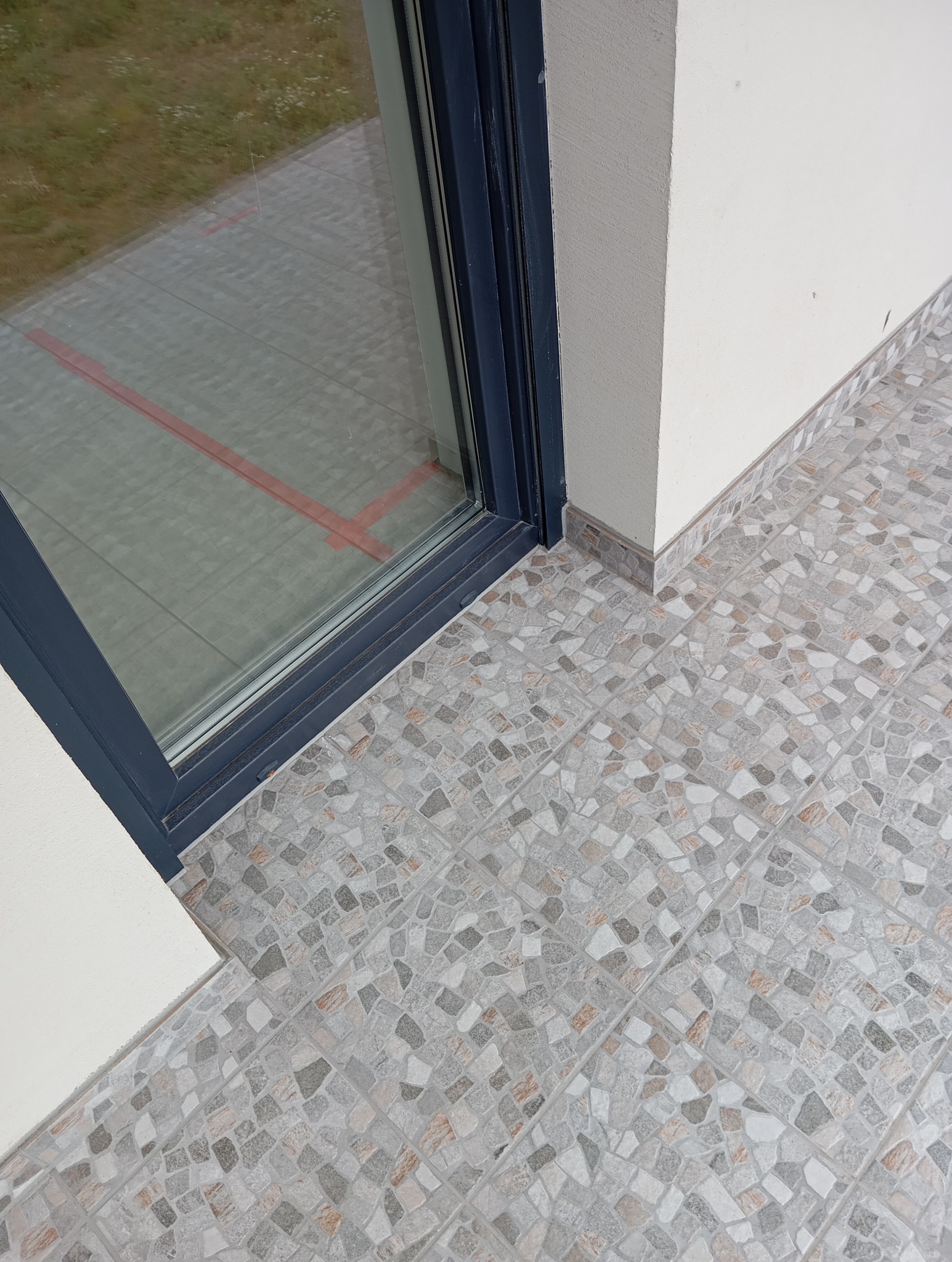 Ułożona mozaika z płytek w odcieniach szarości i beżu na balkonie, widok z góry, z fragmentem szarej ramy okiennej i białej ściany. Drobne elementy tworzą wzór.