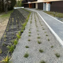 Nasadzenia trawy ozdobnej na osiedlu Villa Park w Brzesku, widok na fragment ogrodu z czarną agrowłókniną i szarym żwirem, na tle nowoczesny budynek mieszkalny z balkonami.