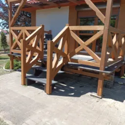 Zadaszona konstrukcja z jasnego drewna z balustradą o wzorze krzyżowym, prowadząca do wejścia domu z widocznymi roletami zewnętrznymi i czerwonym dachem.