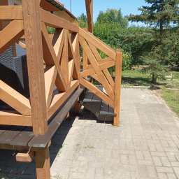 Dariusz Wyrwa - Drewniana konstrukcja z balustradą o geometrycznym wzorze, prowadząca do domu, z widocznymi schodami i elementami pergoli, na tle zieleni i brukowanego chodnika.