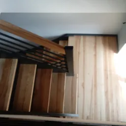 Widok z góry na jasne, drewniane stopnie z czarną metalową balustradą i drewnianą poręczą, oświetlone wpadającym światłem z okna.