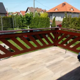 Nowoczesna balustrada z ciemnego drewna na tarasie z płytek imitujących drewno, widok na zadbany ogród z tujami i domy jednorodzinne w tle.