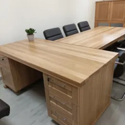 Duży, narożny biurko z jasnego drewna z szufladami, zamykanymi na klucz, w tle trzy krzesła biurowe i szafka, na blacie biurka doniczka z rośliną.