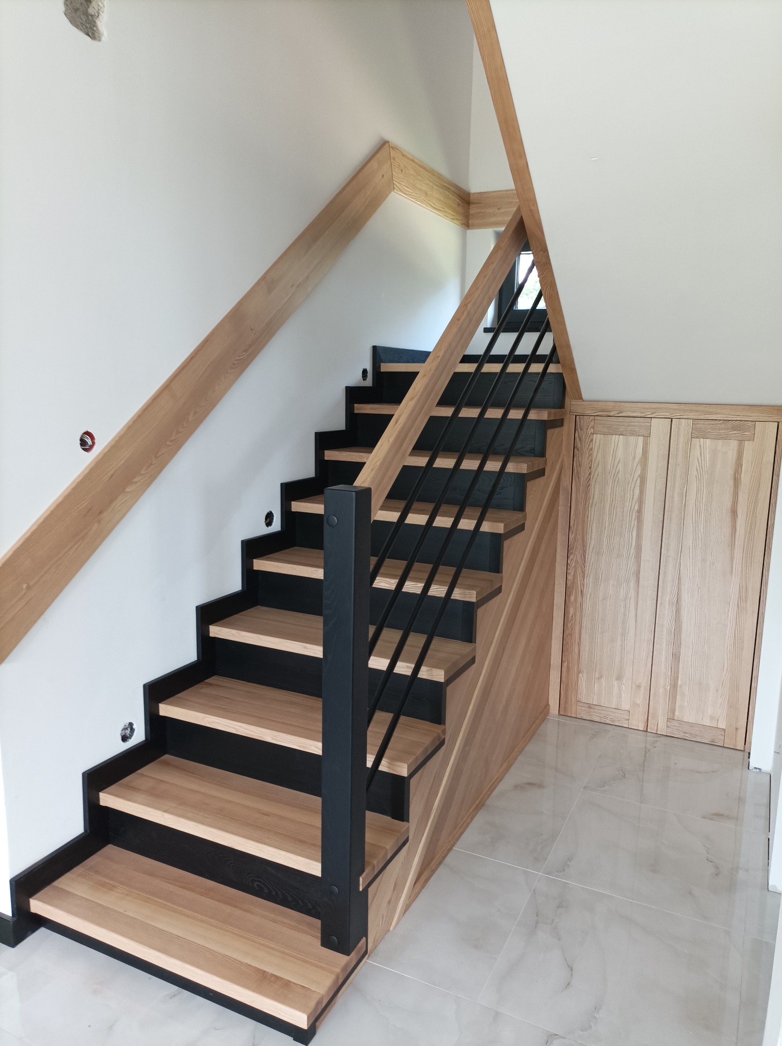 Nowoczesne schody z drewnianymi stopniami i czarnymi podstopnicami oraz metalową balustradą, z drewnianym wykończeniem i zabudową wnęki pod schodami.