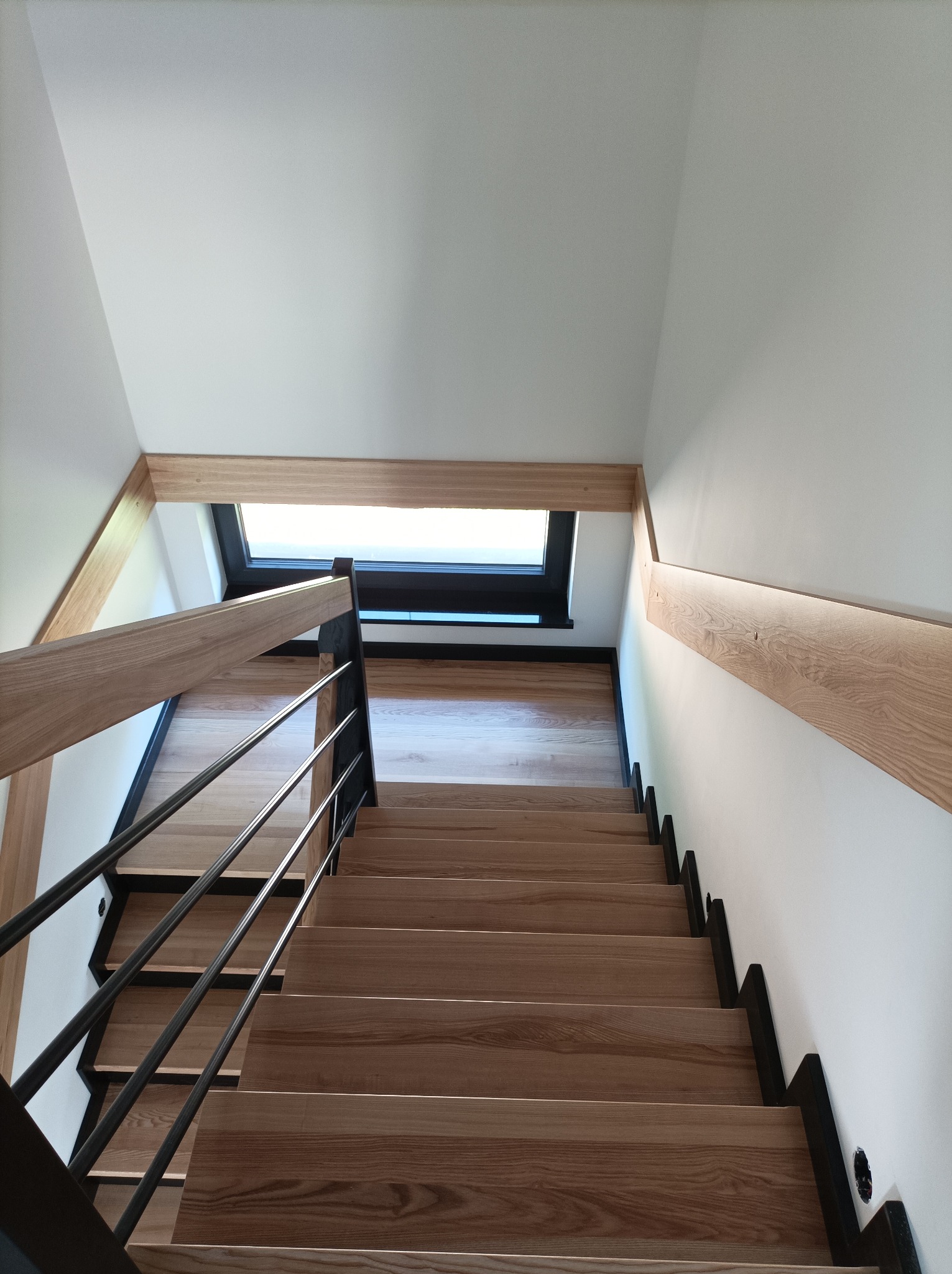 Wnętrze domu z perspektywy schodów z jasnego drewna z czarnymi podstopnicami i metalową balustradą, widok w górę w kierunku okna dachowego.