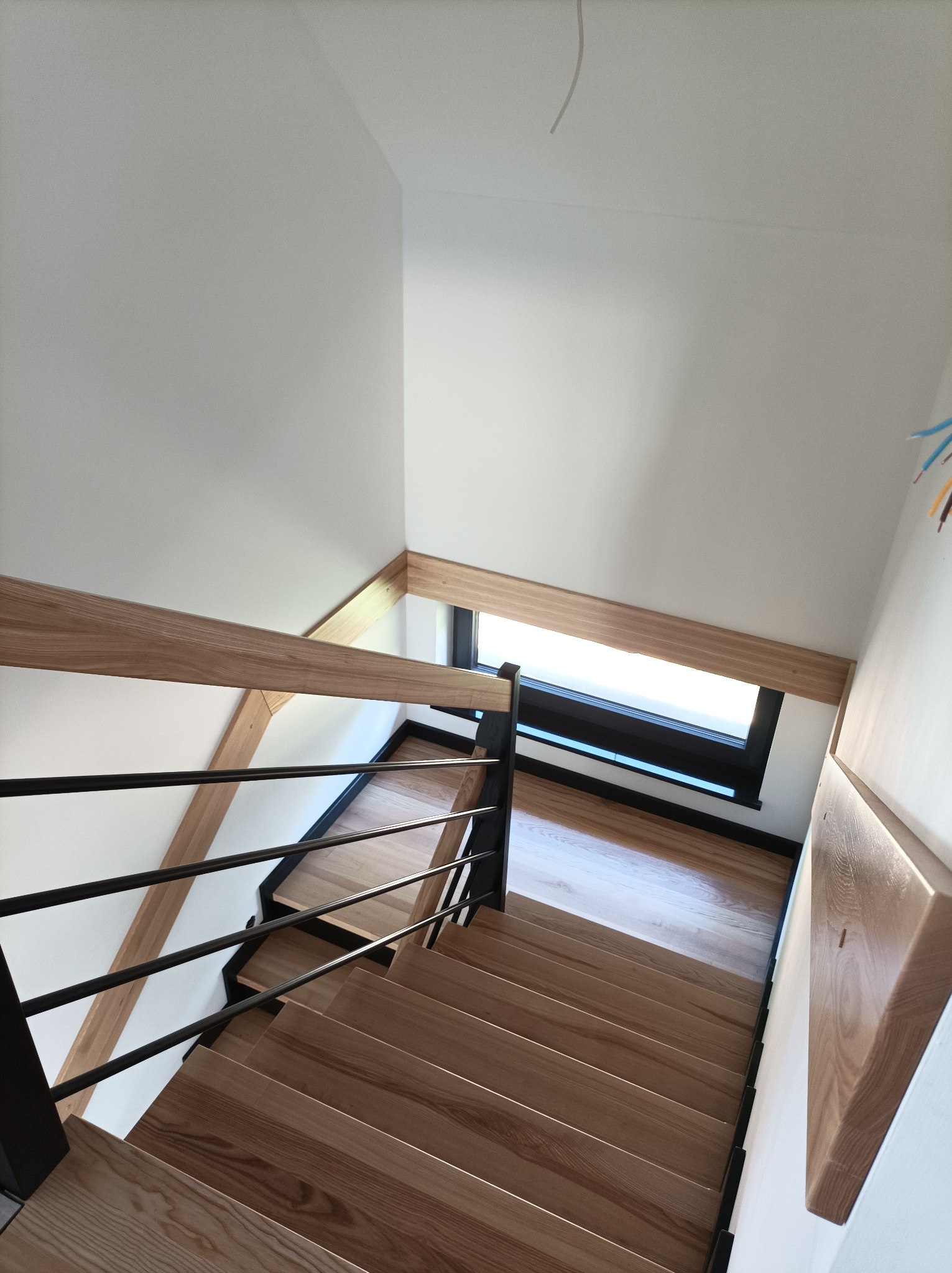 Kątowe ujęcie nowoczesnych schodów z jasnego drewna z czarnymi metalowymi balustradami w jasnym wnętrzu, widoczne okno w ramie i fragment drewnianej półki na ścianie.