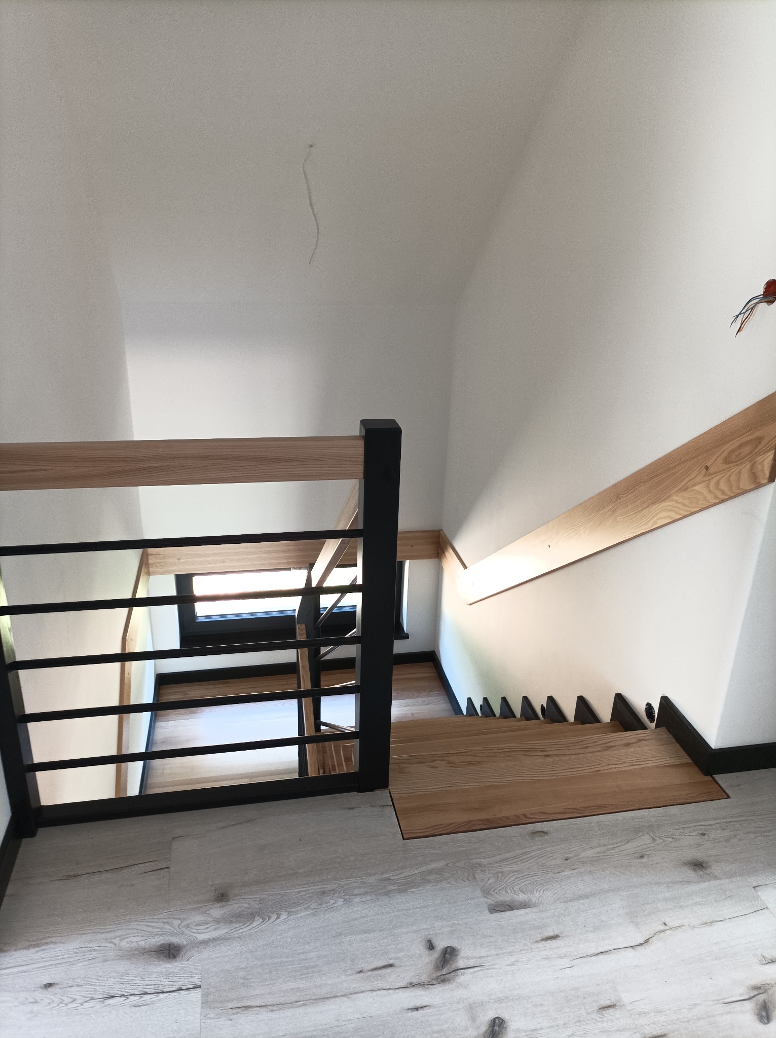 Wnętrze domu z widokiem na dół schodów z drewnianymi stopniami i czarną metalową balustradą z drewnianą poręczą, widoczne niedokończone wykończenie ściany z wystającymi przewodami.