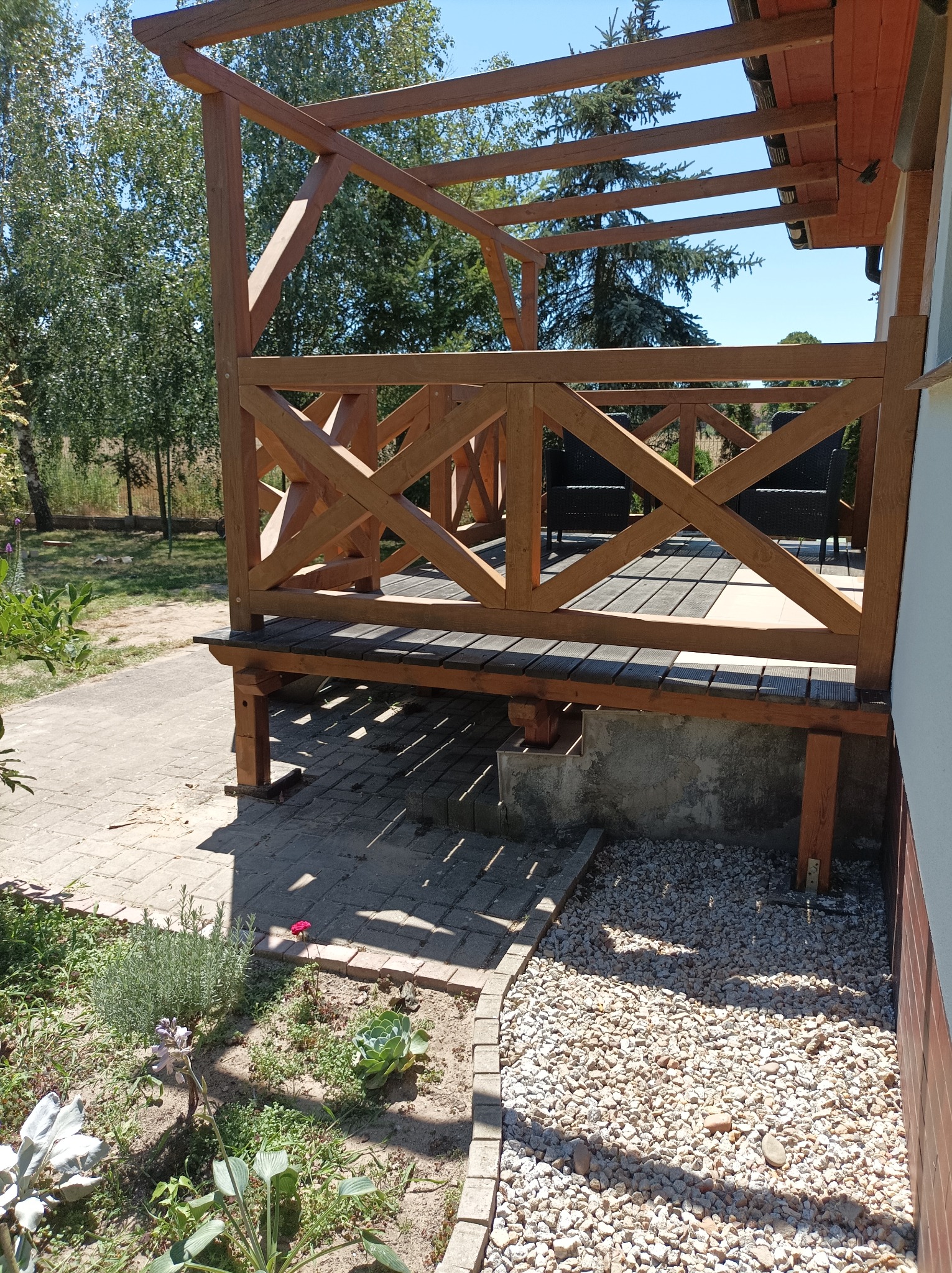 Drewniana konstrukcja z balustradą o wzorze krzyżowym, tworząca zadaszone patio przy domu, widok z zewnątrz na fragment ogrodu z roślinami i kamieniami.