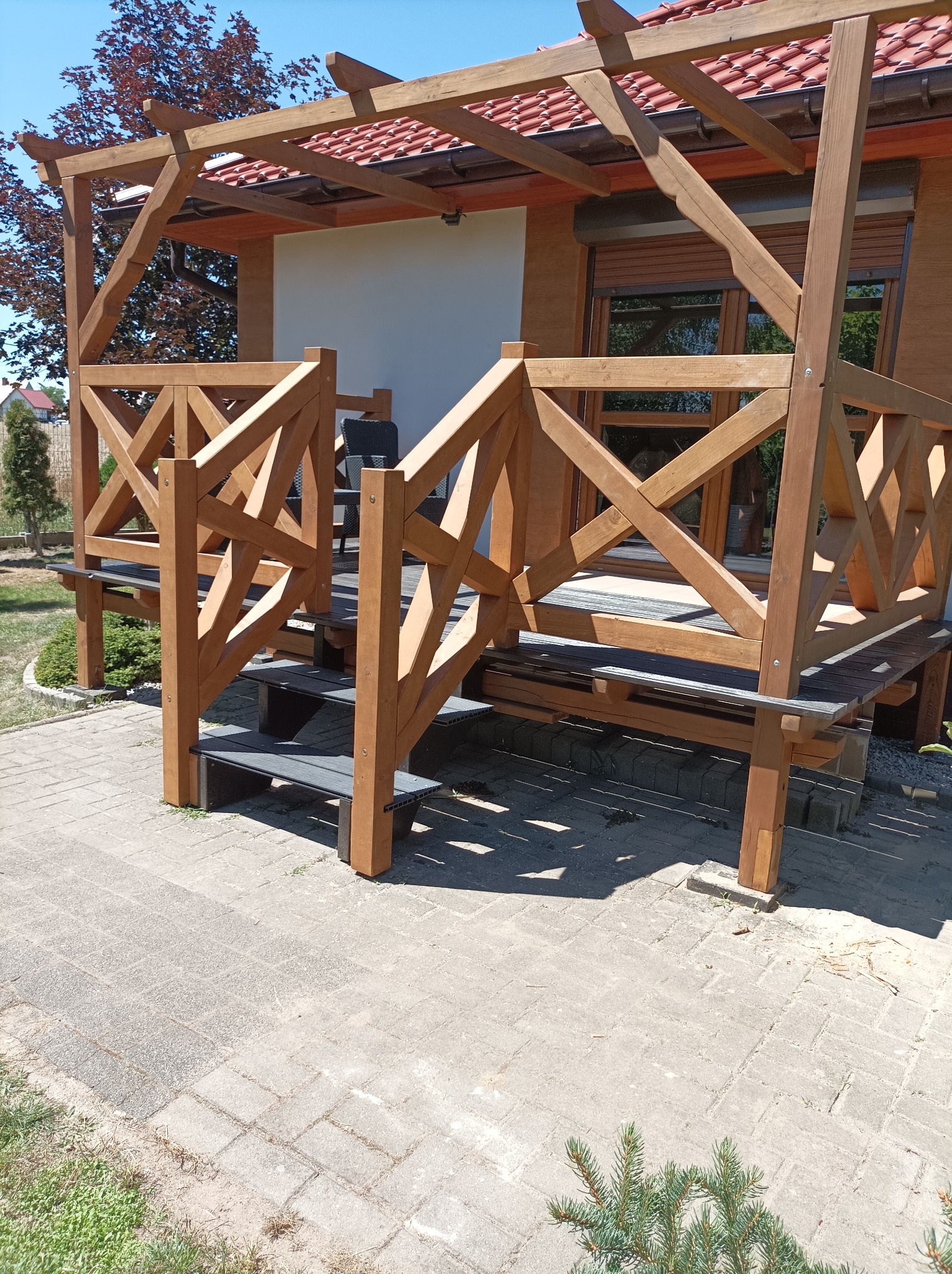 Zadaszona konstrukcja z jasnego drewna z balustradą o wzorze krzyżowym, prowadząca do wejścia domu z widocznymi roletami zewnętrznymi i czerwonym dachem.