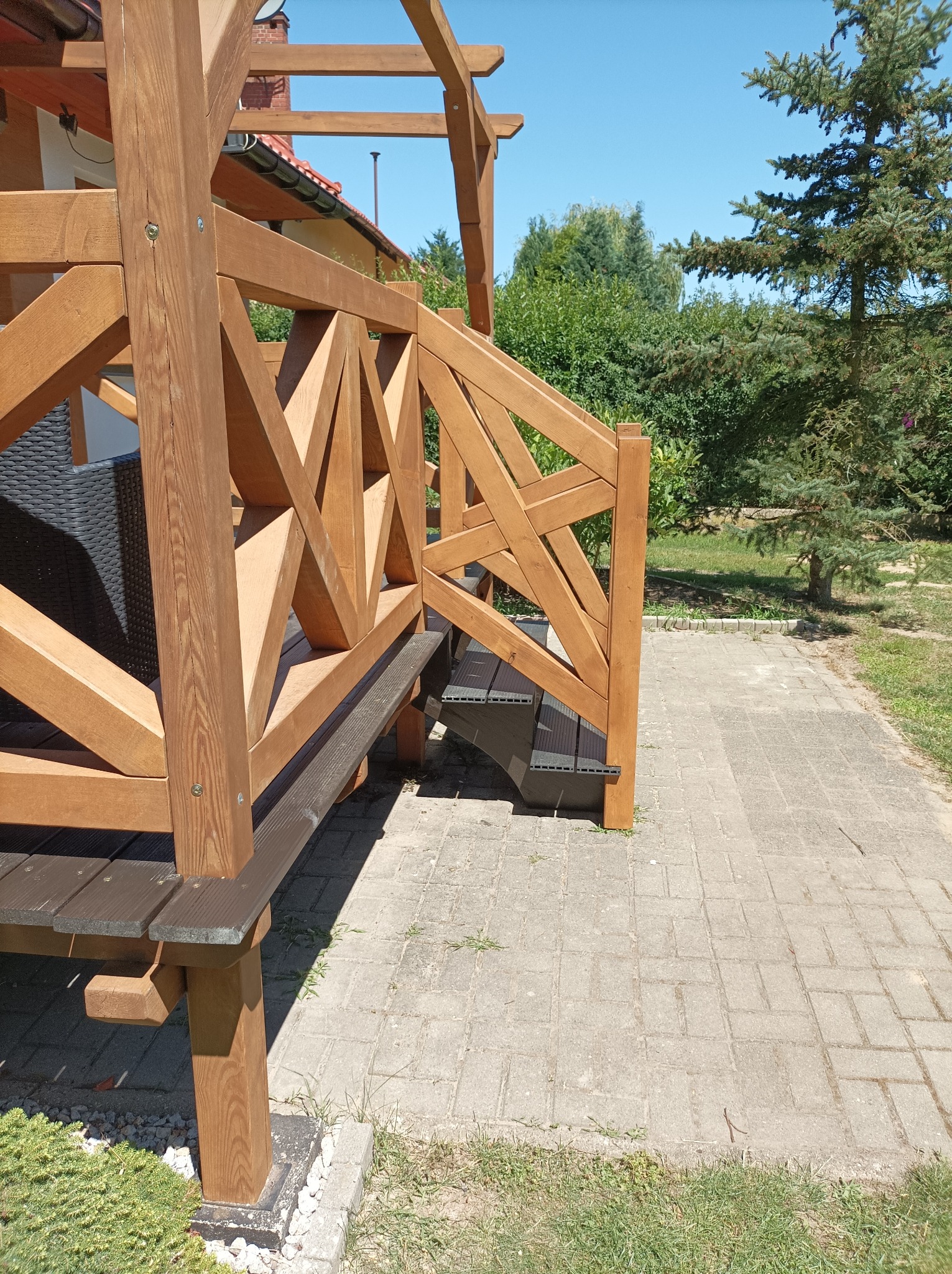 Drewniana konstrukcja z balustradą o geometrycznym wzorze, prowadząca do domu, z widocznymi schodami i elementami pergoli, na tle zieleni i brukowanego chodnika.