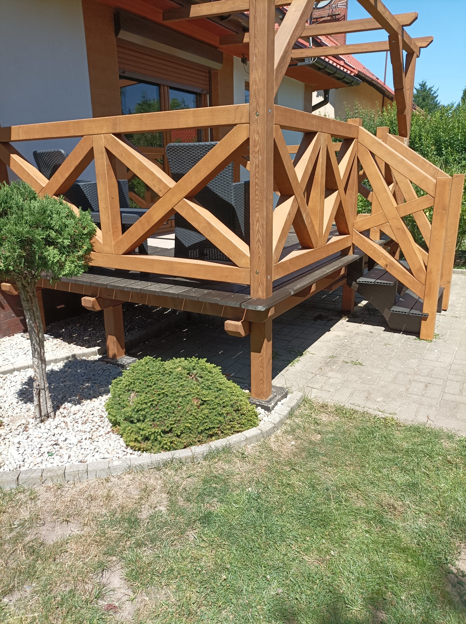 Drewniana konstrukcja tarasu z balustradą o geometrycznym wzorze krzyży, schody wejściowe, widok na ogród z ozdobnymi krzewami i trawnikiem.