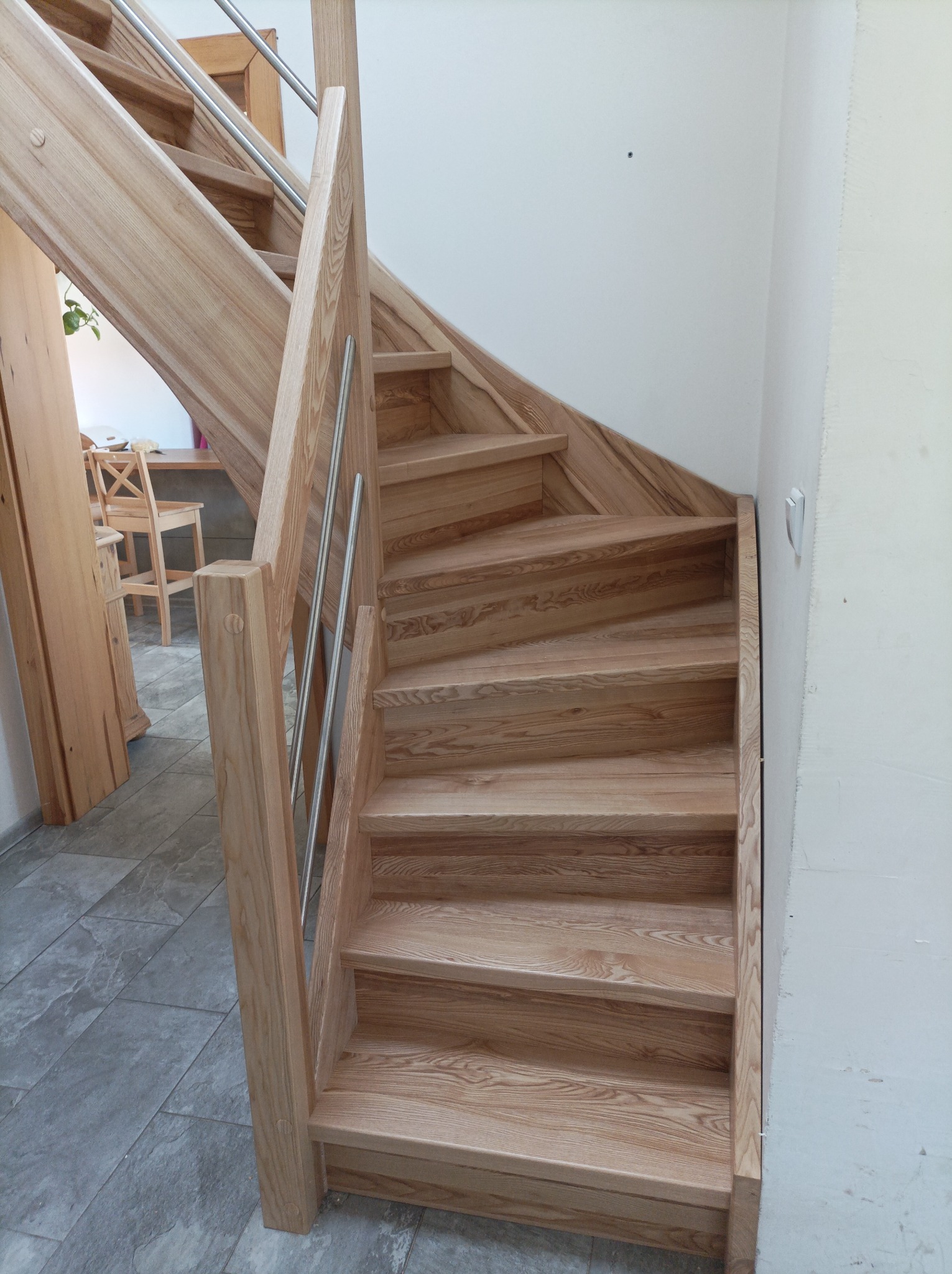 Jasne, drewniane schody z metalową balustradą w nowoczesnym wnętrzu, widoczne fragmenty podłogi z szarych płytek i białej ściany.