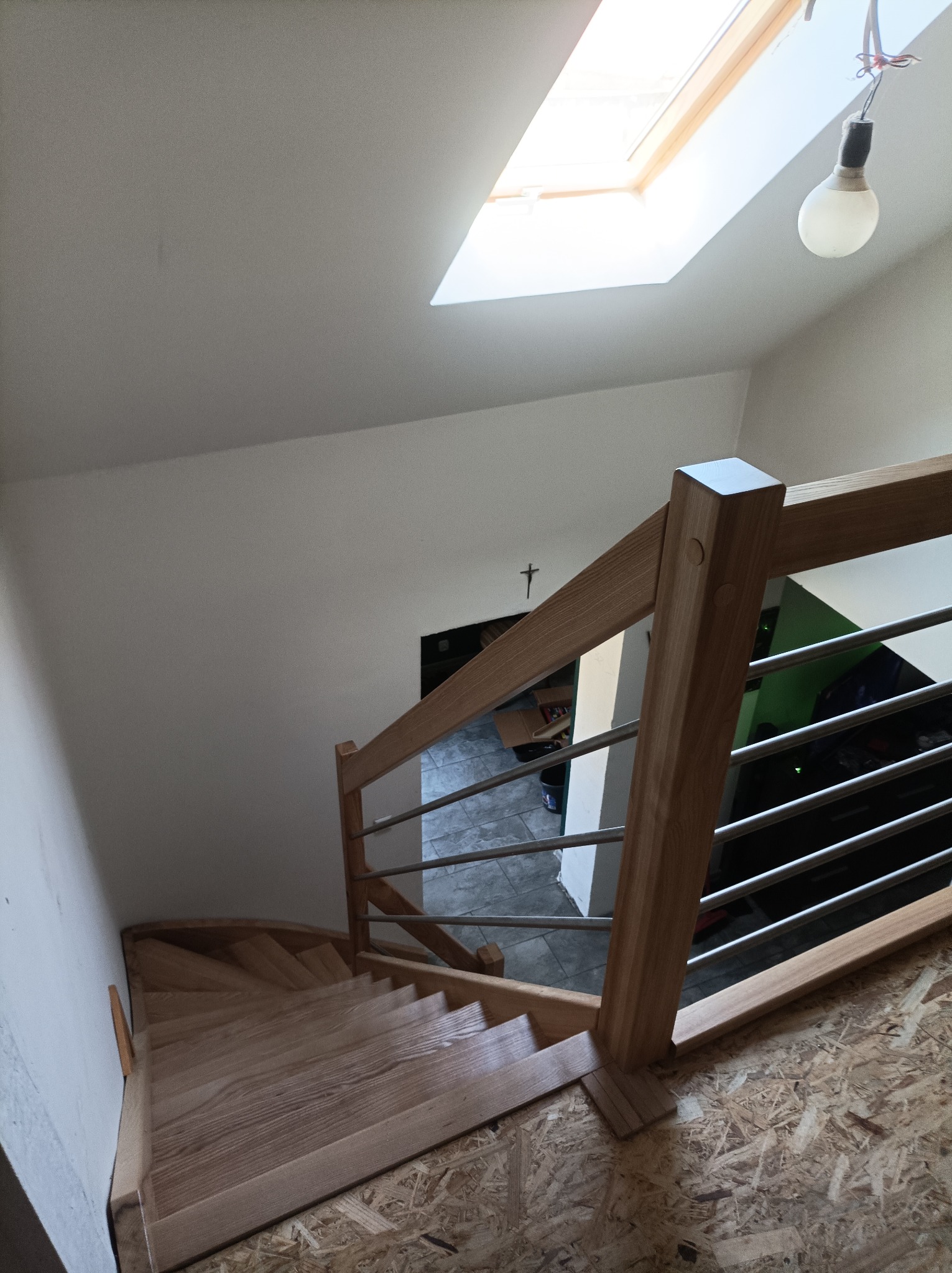 Drewniane schody z balustradą ze stali nierdzewnej, widok z góry na zakręt schodów, surowy stan wykończenia podłogi, oświetlenie naturalne z okna dachowego.