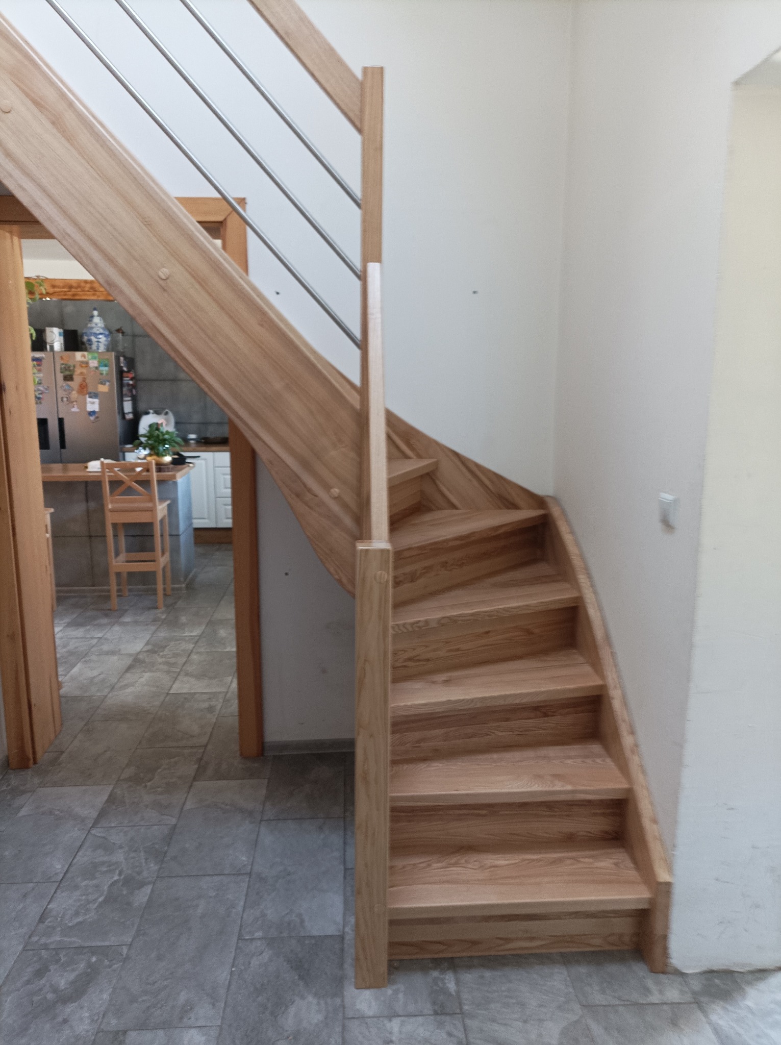 Jasne, drewniane schody z metalową balustradą, skręcające w lewo, widoczne z dołu, z fragmentem kuchni w tle.