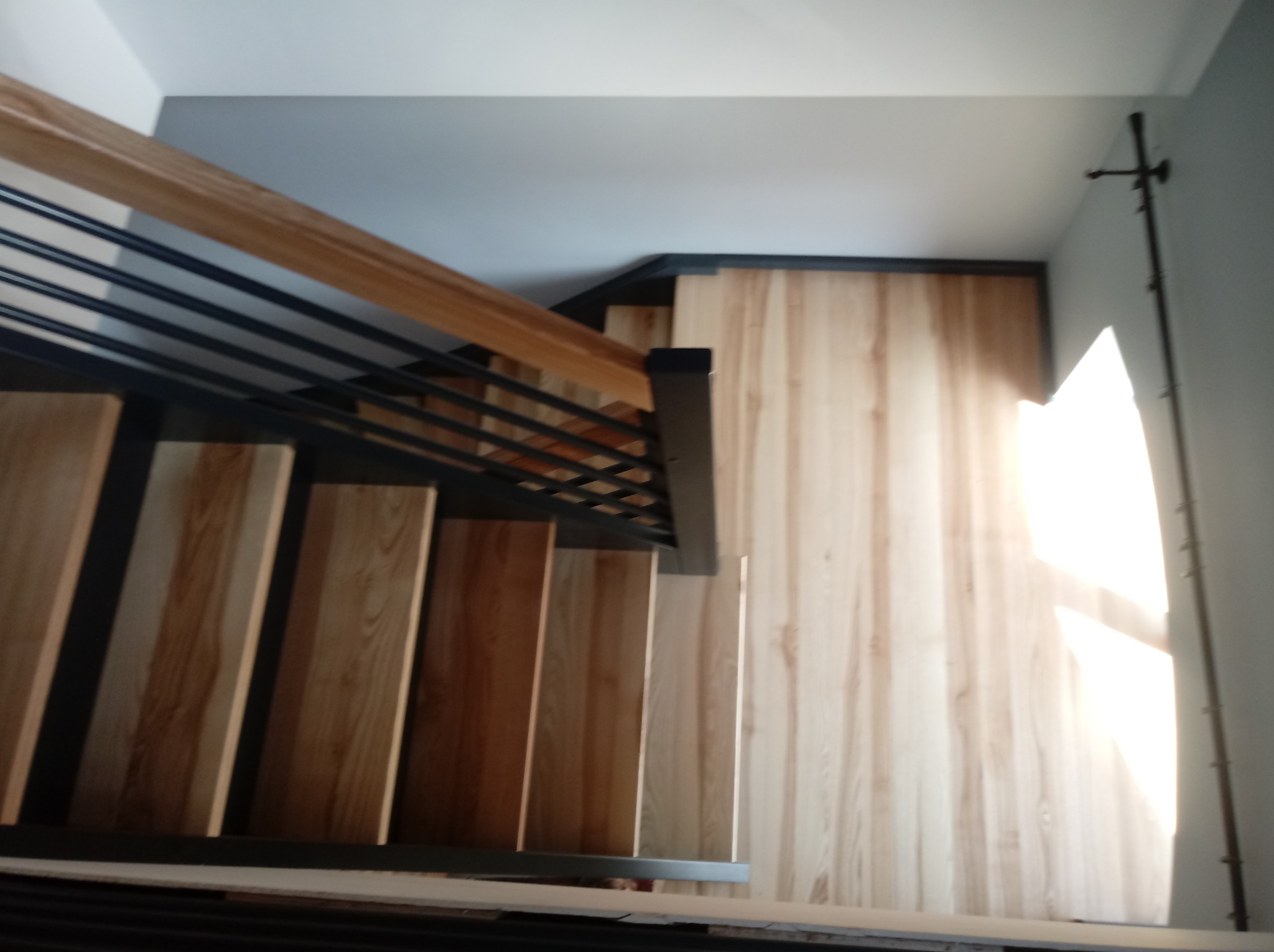 Widok z góry na jasne, drewniane stopnie z czarną metalową balustradą i drewnianą poręczą, oświetlone wpadającym światłem z okna.