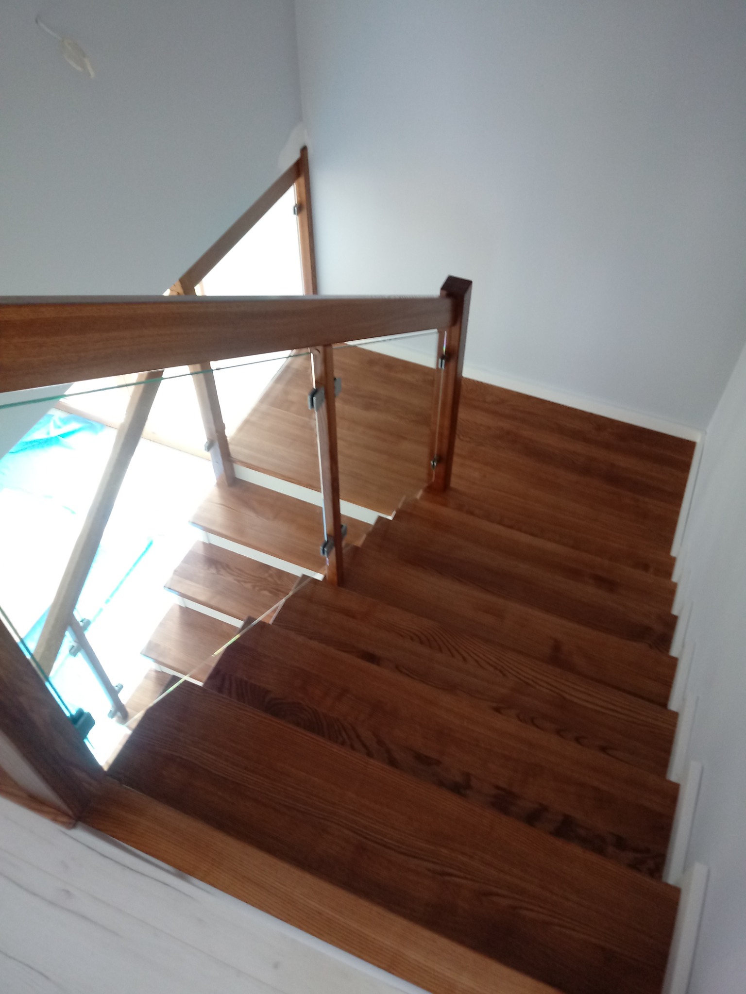 Widok z góry na brązowe stopnie z drewna, z przezroczystą szklaną balustradą i drewnianą poręczą, w jasnym wnętrzu.