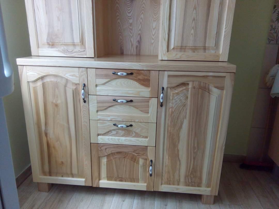 Drewniana komoda z nadstawką, widoczne trzy szuflady z ozdobnymi uchwytami oraz dwoje drzwi z wyfrezowanymi panelami, wykonana z jasnego drewna o wyraźnym usłojeniu.