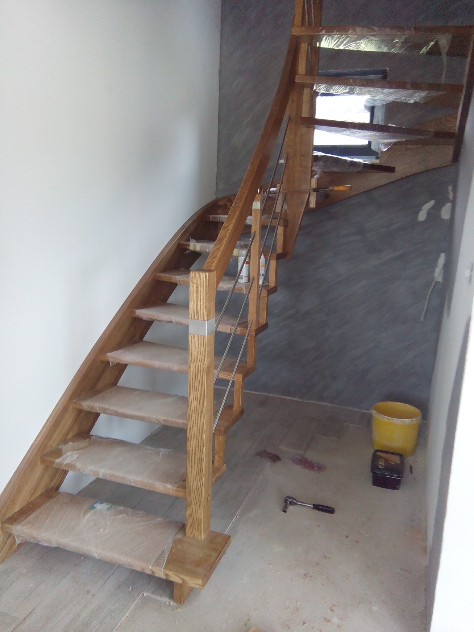 Wewnętrzne, drewniane schody z metalową balustradą w trakcie montażu, zabezpieczone folią, widoczny żółty kubeł i narzędzia na podłodze.