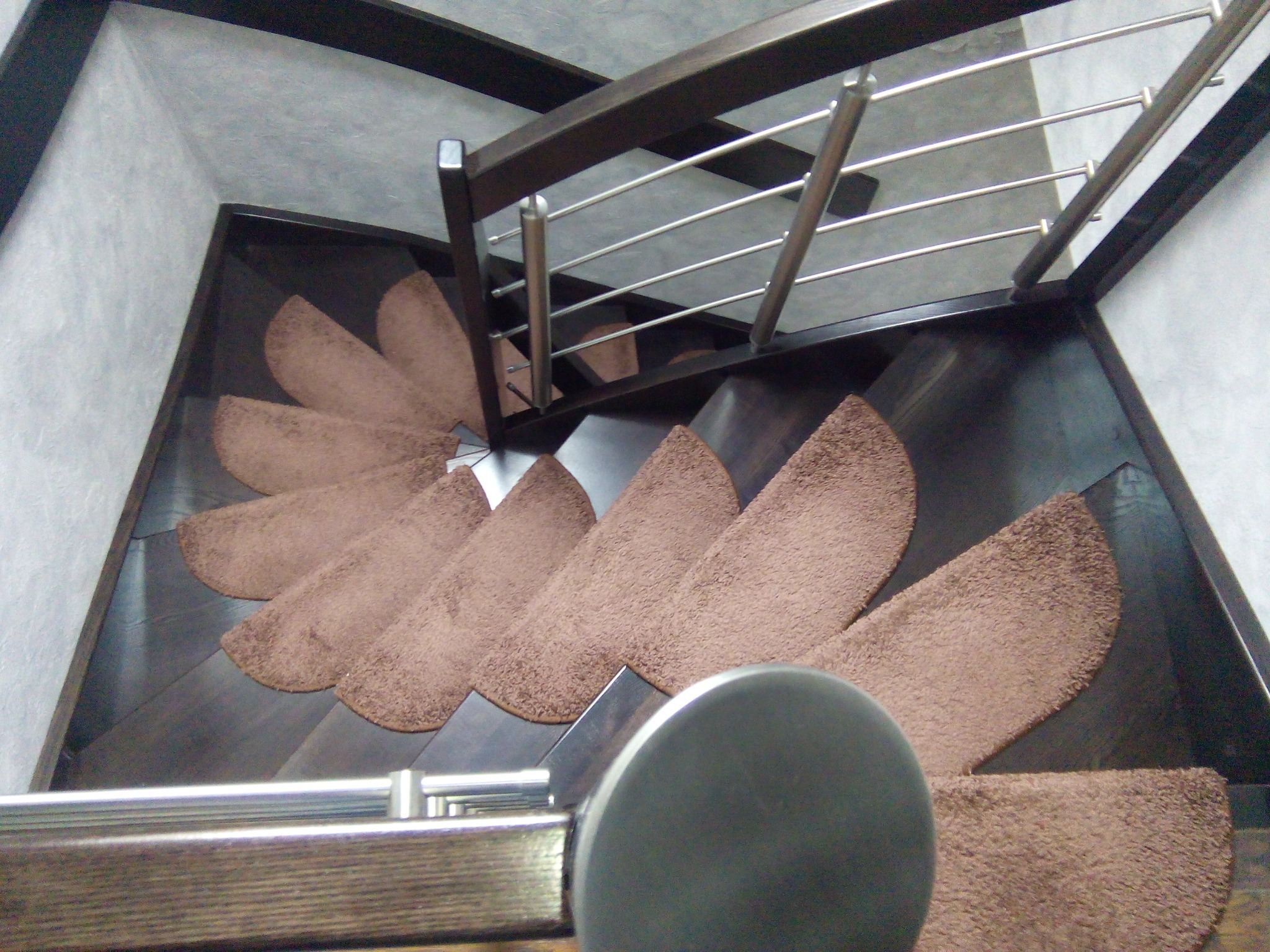 Kątowe ujęcie ciemnych schodów z brązowymi, półokrągłymi nakładkami dywanowymi, zabezpieczonych balustradą z drewnianą ramą i metalowymi prętami.