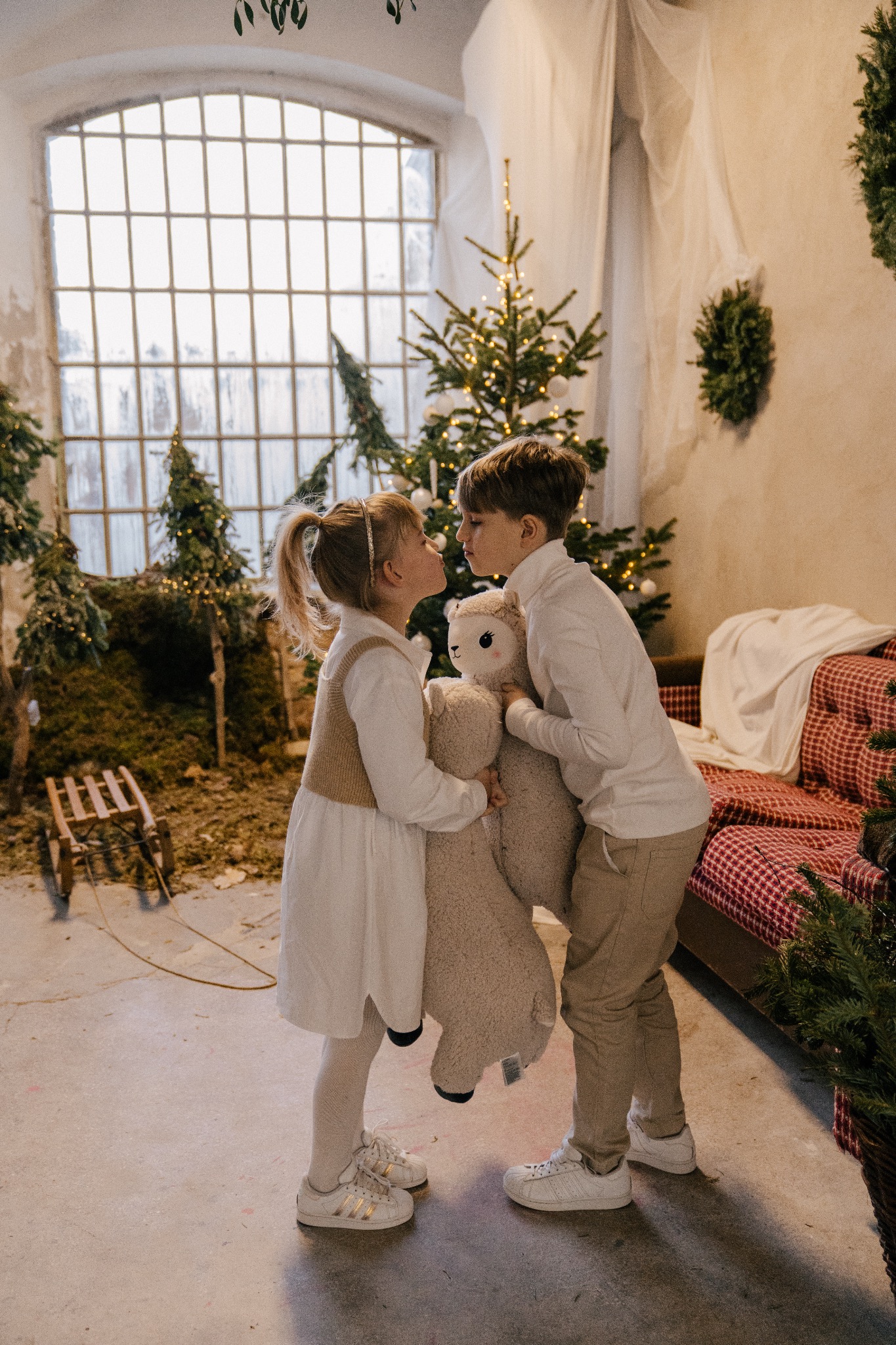 Dzieci trzymające pluszową lamę w świątecznej scenerii z choinkami i dużym oknem, zbliżają się do siebie twarzami.