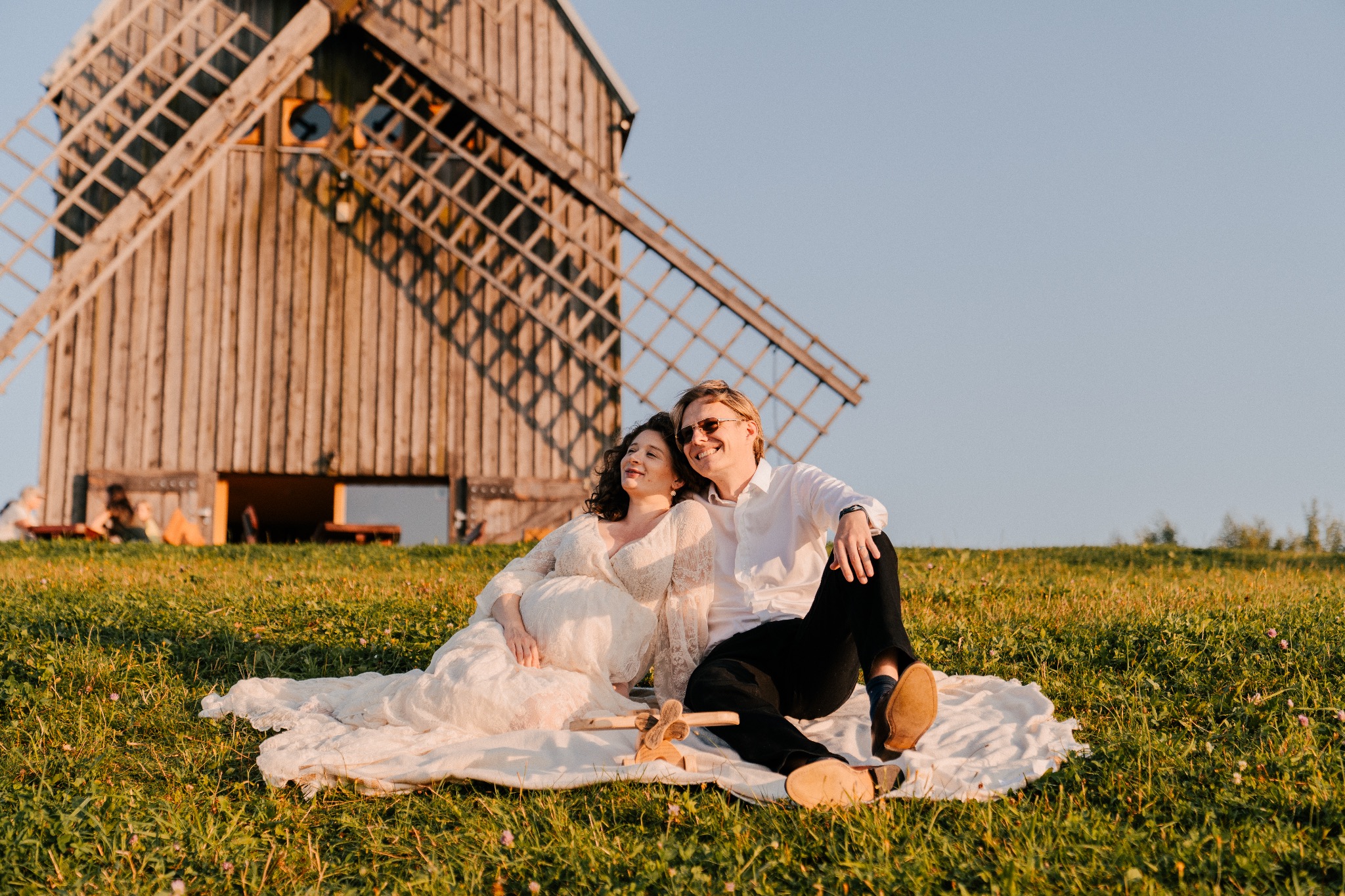 Uśmiechnięta para w oczekiwaniu na dziecko, siedząca na kocu na zielonej łące z drewnianym wiatrakiem w tle, trzymająca drewnianą zabawkę.