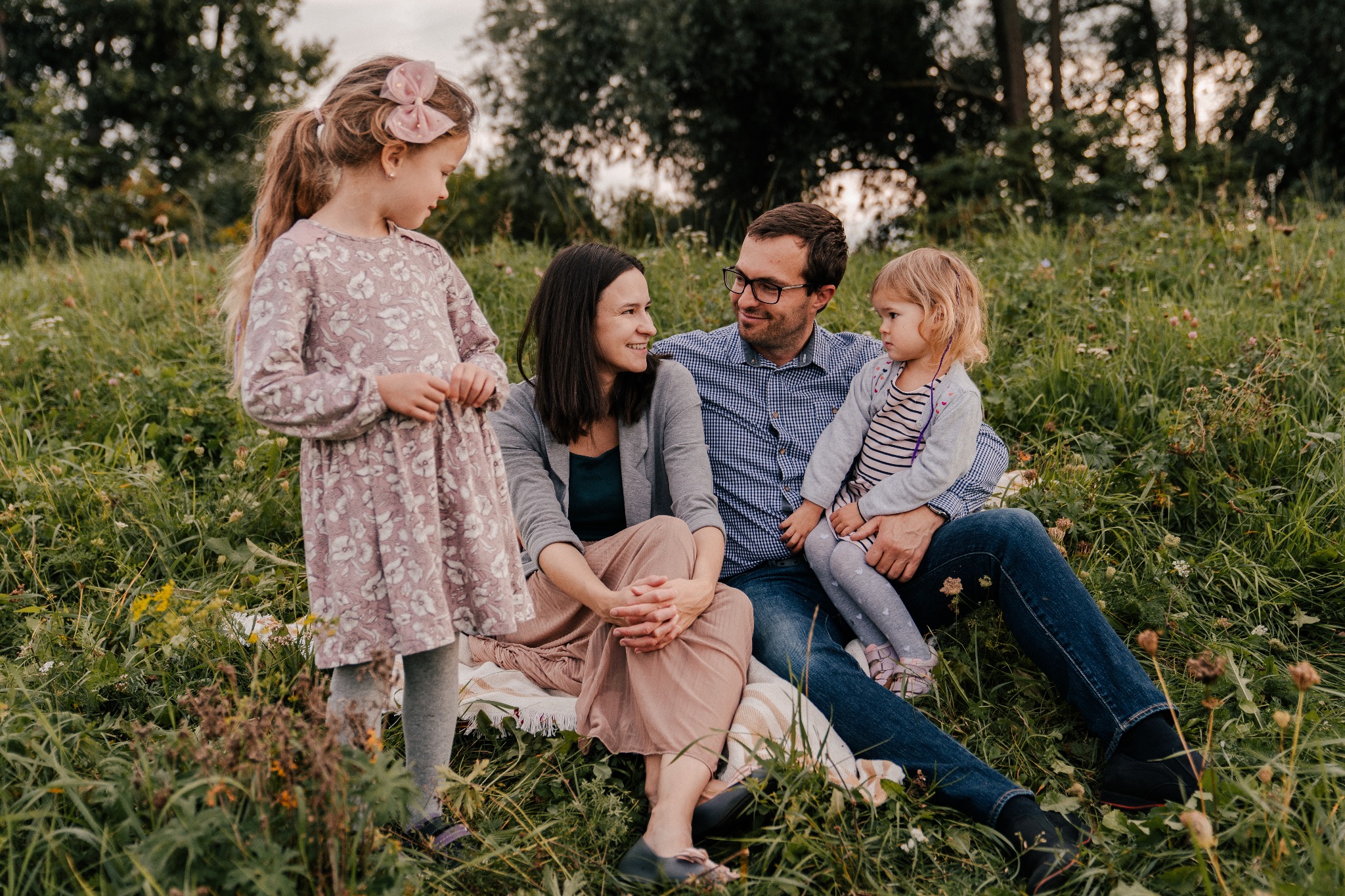 Rodzina z dwiema córkami siedzi na kocu w wysokiej trawie podczas sesji zdjęciowej, starsza córka ma różową kokardę we włosach.