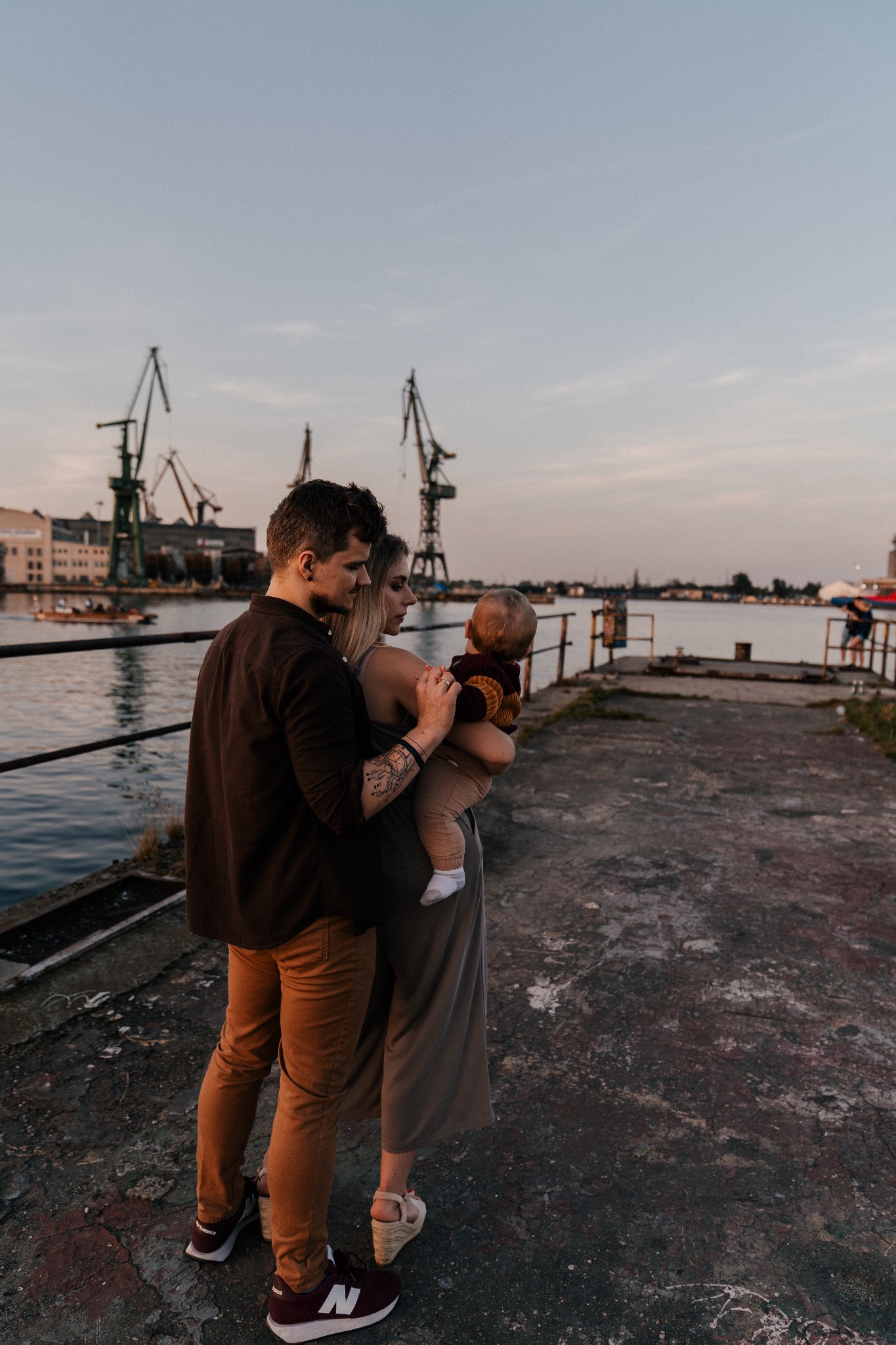 Rodzina na tle portowych żurawi: ojciec obejmuje matkę trzymającą dziecko, widok z boku na nabrzeże i wodę.