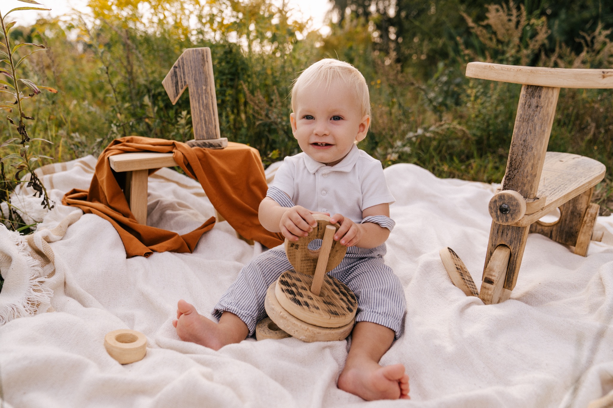 Radosny roczny chłopiec siedzi na białym kocu w plenerze, trzymając drewnianą zabawkę, obok drewniana jedynka i miniaturowy drewniany fotel.