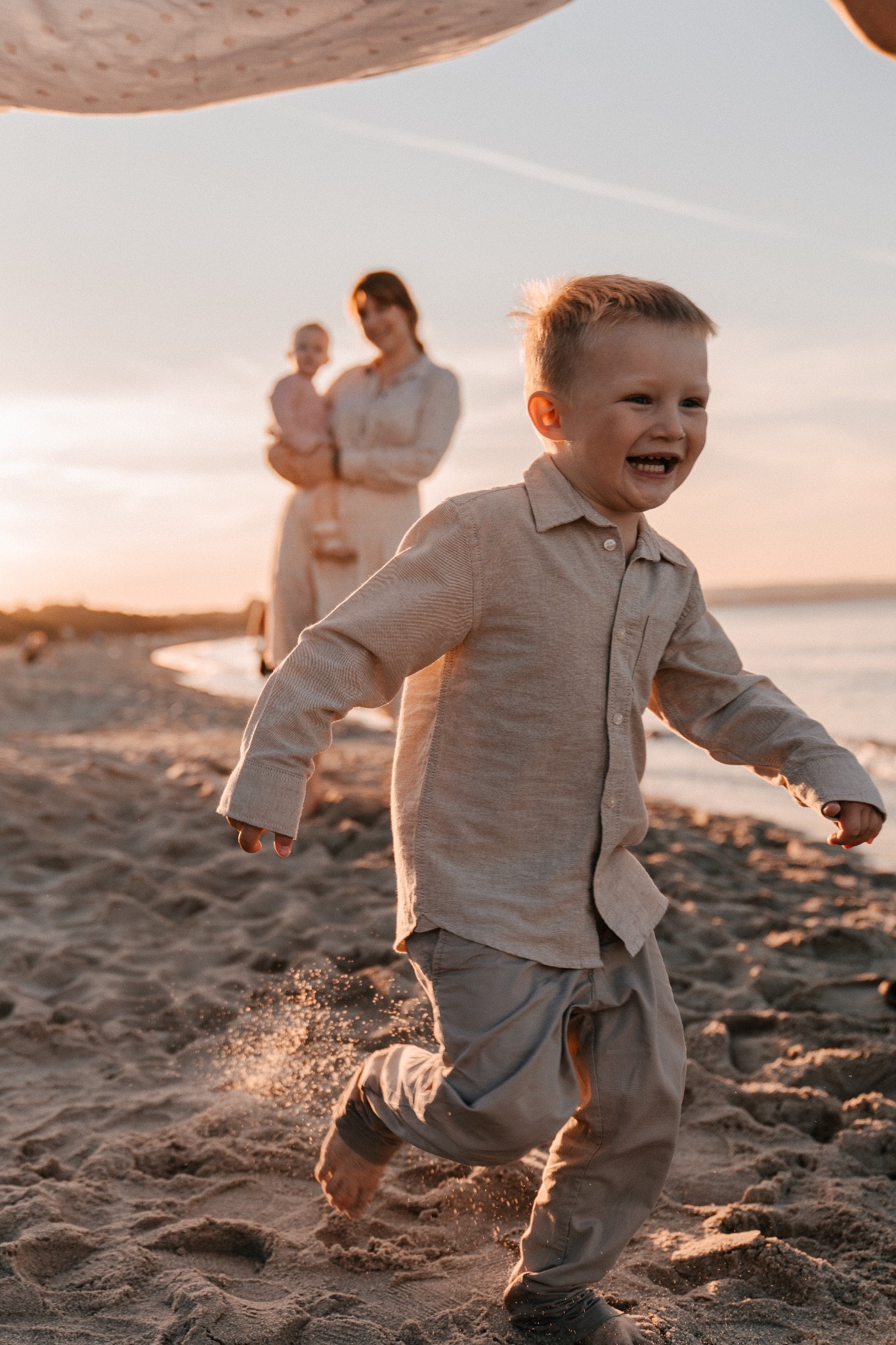 Uśmiechnięty chłopiec biegnie boso po piaszczystej plaży w promieniach zachodzącego słońca, w tle matka trzymająca niemowlę.
