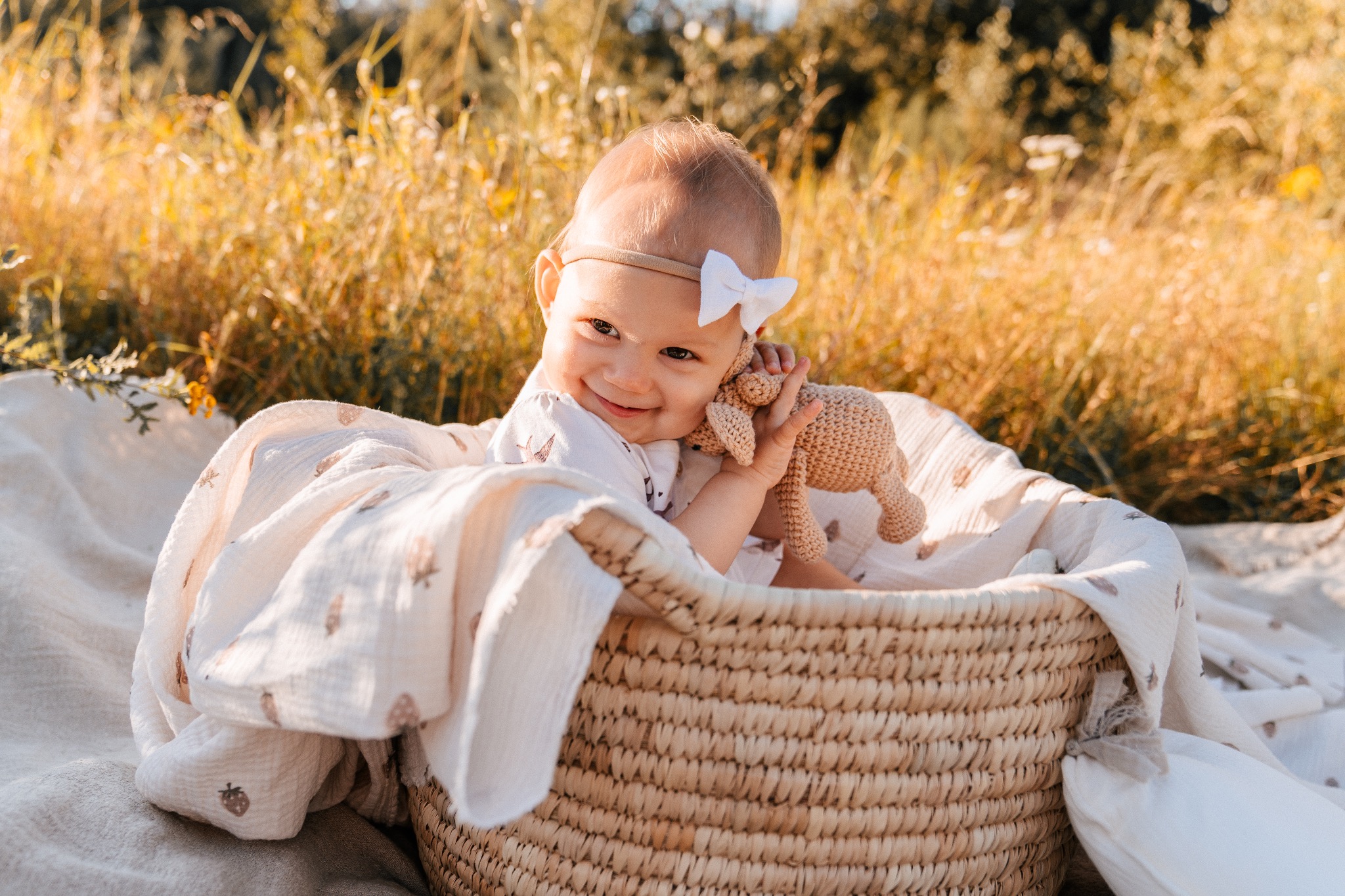 Uśmiechnięta niemowlęca dziewczynka z białą kokardką na głowie, siedząca w wiklinowym koszu na tle rozświetlonej słońcem trawy, trzymająca pluszowego króliczka.