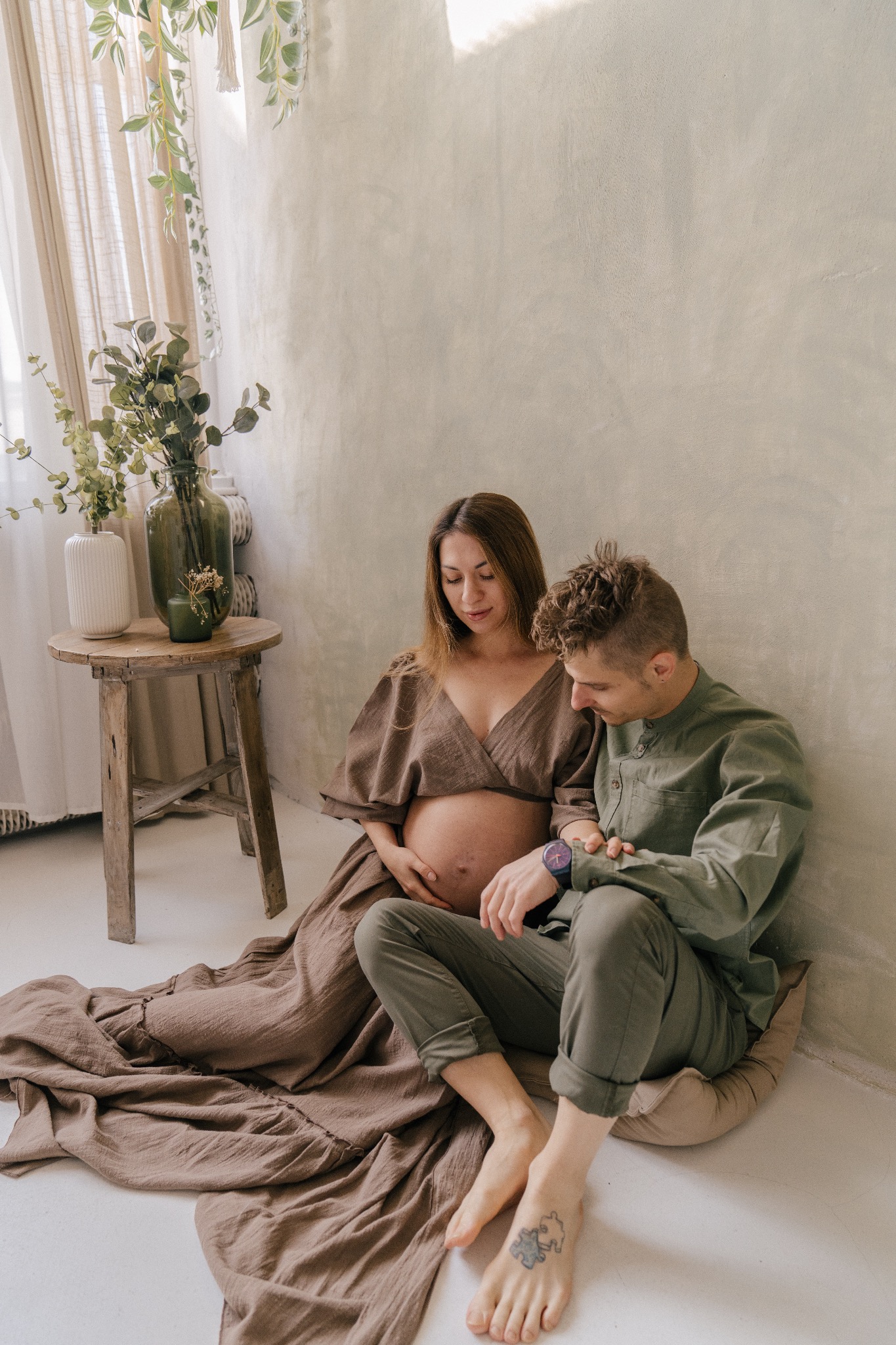 Przyszła mama w brązowej sukience i przyszły tata w zielonej koszuli siedzą na podłodze w minimalistycznym studio, kobieta trzyma dłoń na brzuchu, mężczyzna patrzy na jej ciążowy brzuch, na stopie...