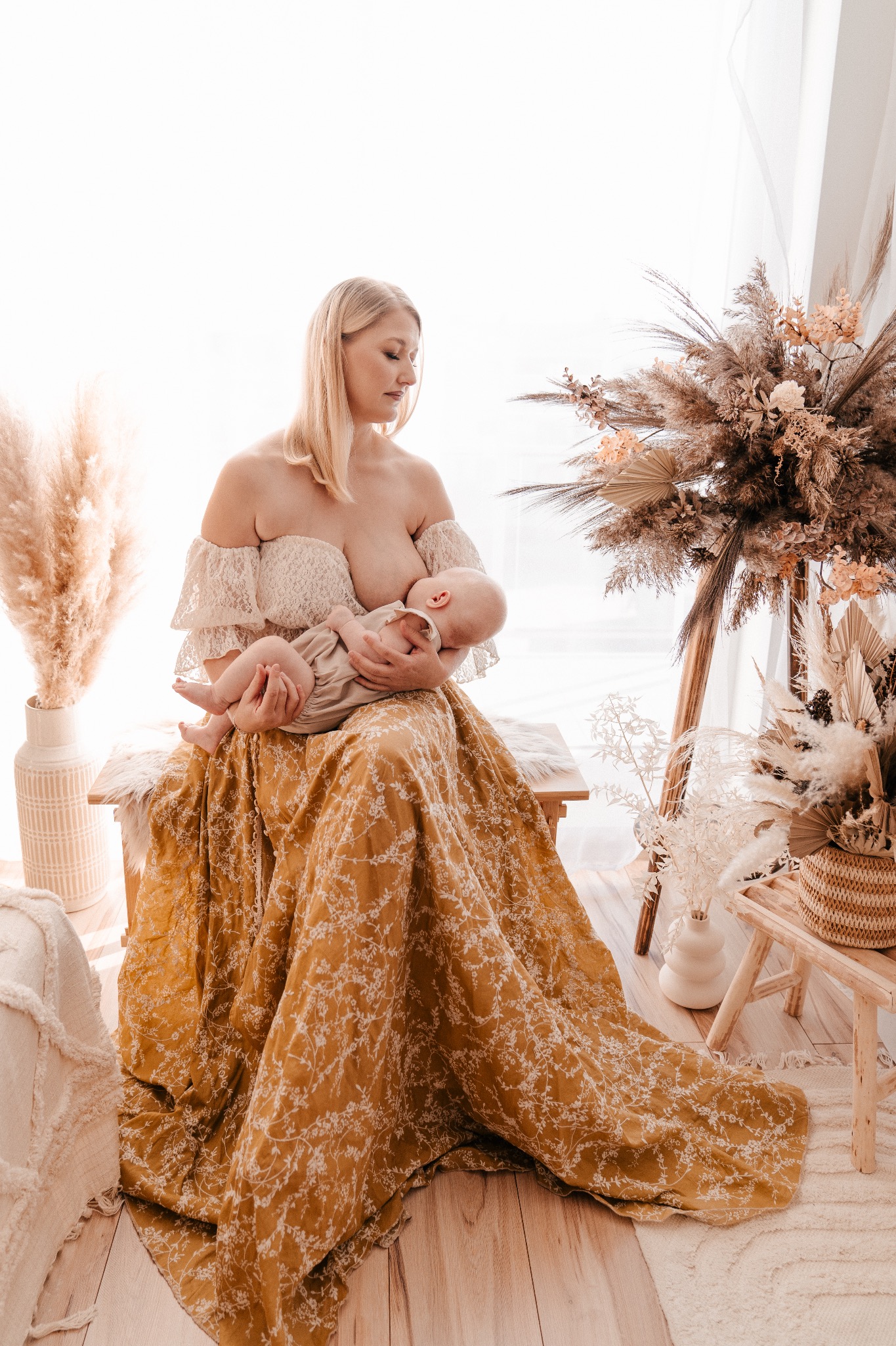 Matka karmiąca noworodka piersią, siedząca na drewnianej ławce w studiu z dekoracjami z suszonych traw i kwiatów w odcieniach beżu i brązu, ubrana w suknię w kolorze musztardowym z białym kwiatowym...