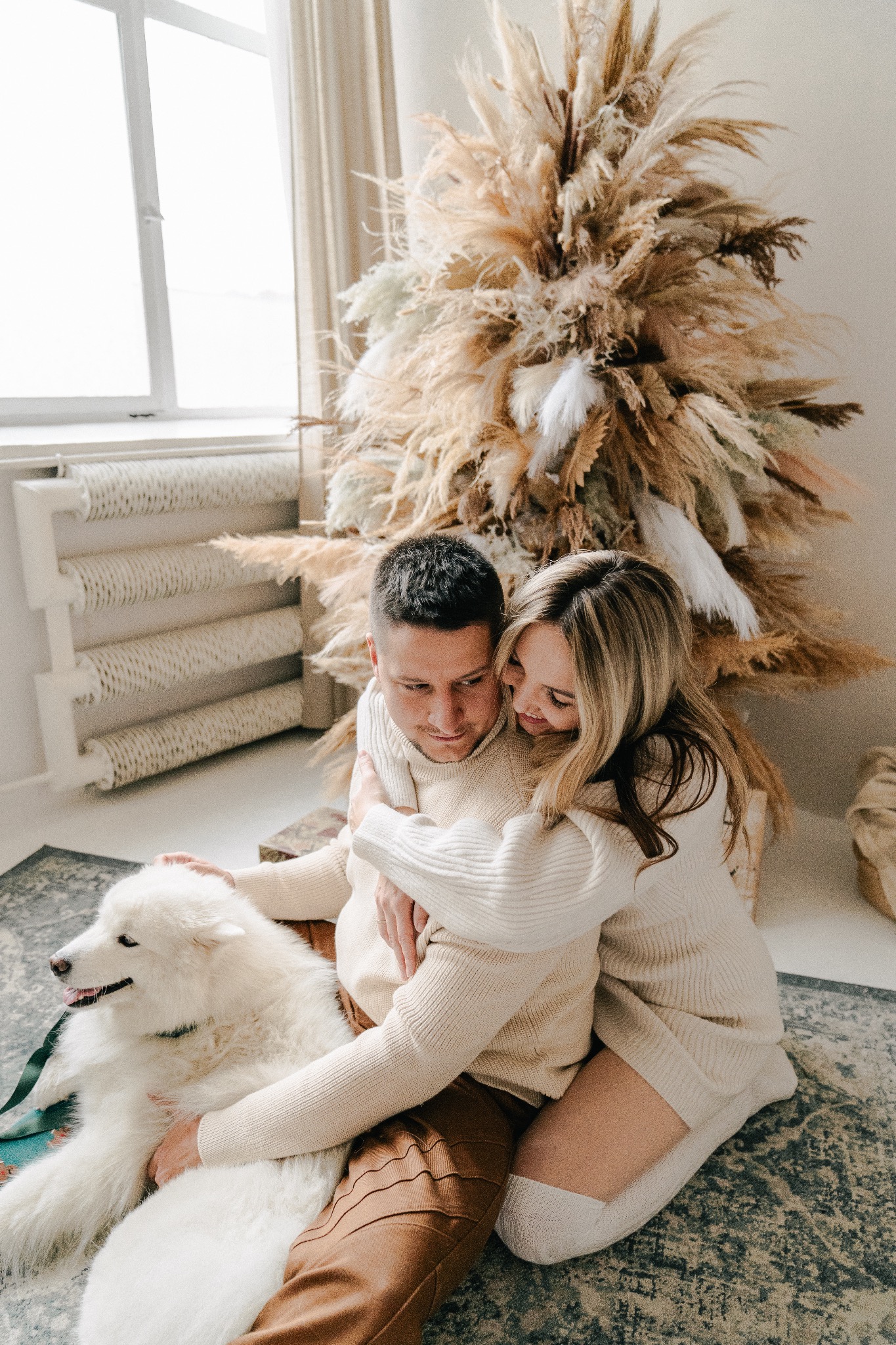 Para w beżowych swetrach obejmuje się siedząc na dywanie obok białego psa Samoyeda, z dekoracją z suszonych traw w tle.