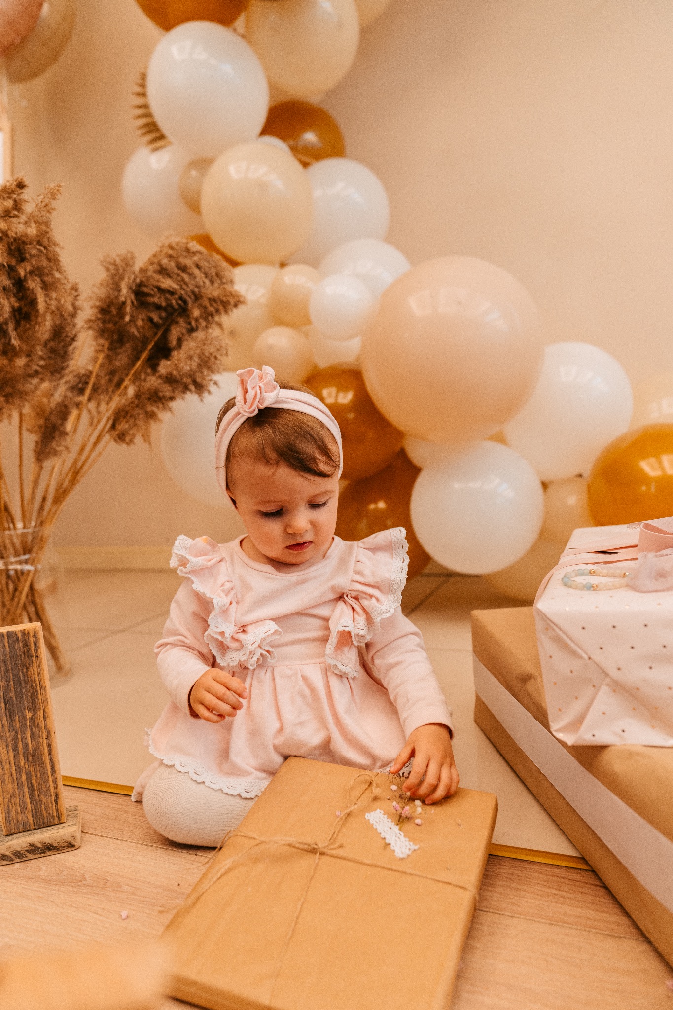 Dziewczynka w różowej sukience i opasce siedzi na podłodze, dotykając prezentu z jutowym sznurkiem i suszonymi kwiatami, w tle balony w odcieniach beżu i brązu.