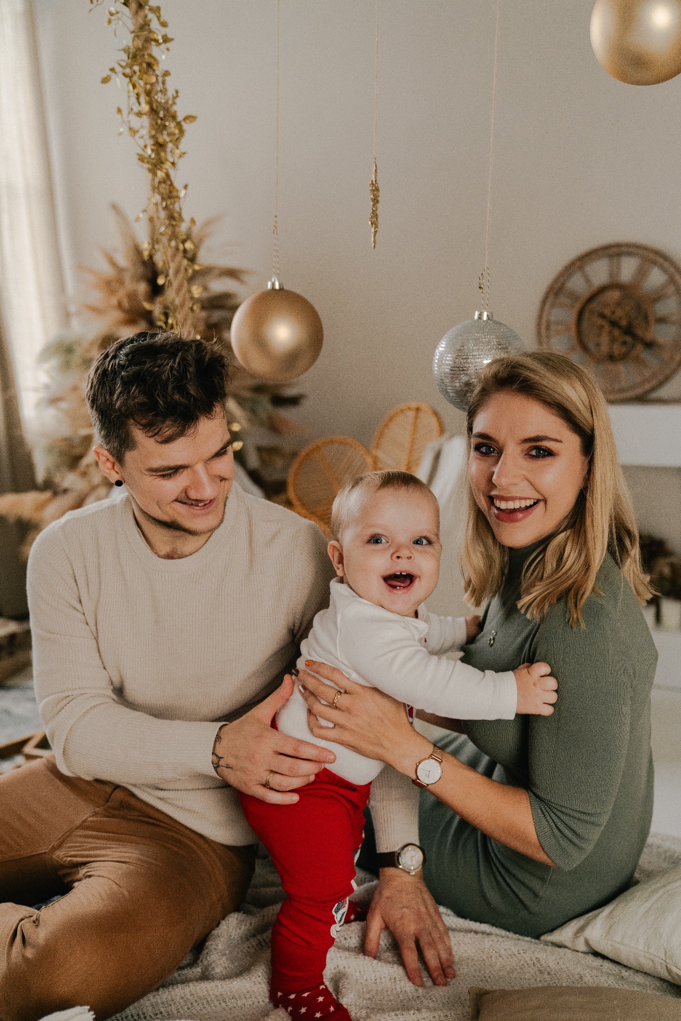 Uśmiechnięta rodzina z małym dzieckiem w czerwonych spodniach na tle dekoracji świątecznych z wiszącymi złotymi kulami i zegarem.