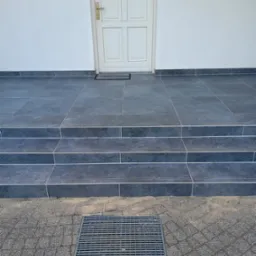 Wykończone płytkami schody wejściowe do domu z białymi drzwiami i kratką odpływową. Ciemnoszare płytki z jasnymi fugami. Widok na elewację i chodnik z kostki.