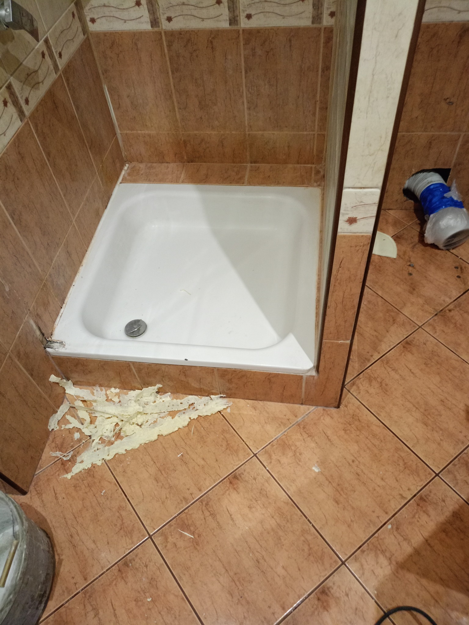 Niedokończona instalacja brodzika w łazience z brązowymi płytkami, widoczne pozostałości pianki montażowej na podłodze.
